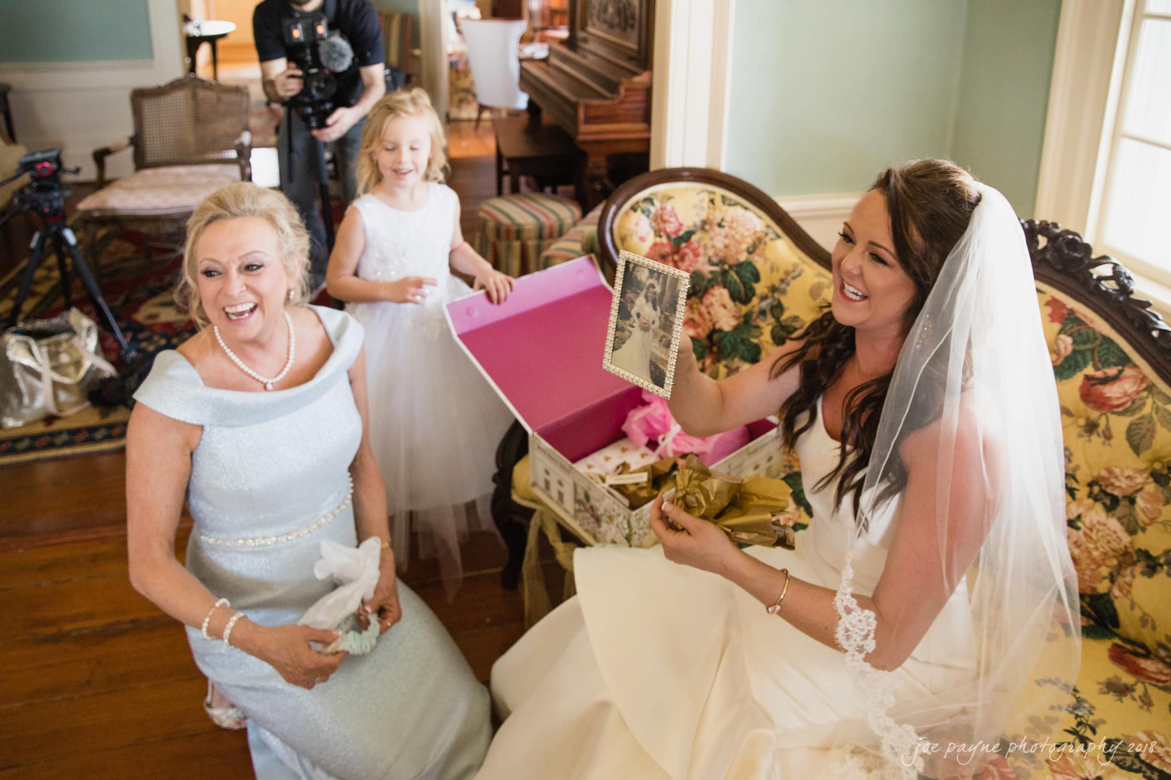 may-lew farm wedding photography - macy & jordan A bride in a white wedding dress and veil joyfully opens a gift, sitting on a vintage sofa. A smiling woman in a light blue dress and a young girl in a white dress stand nearby in a warmly decorated room.