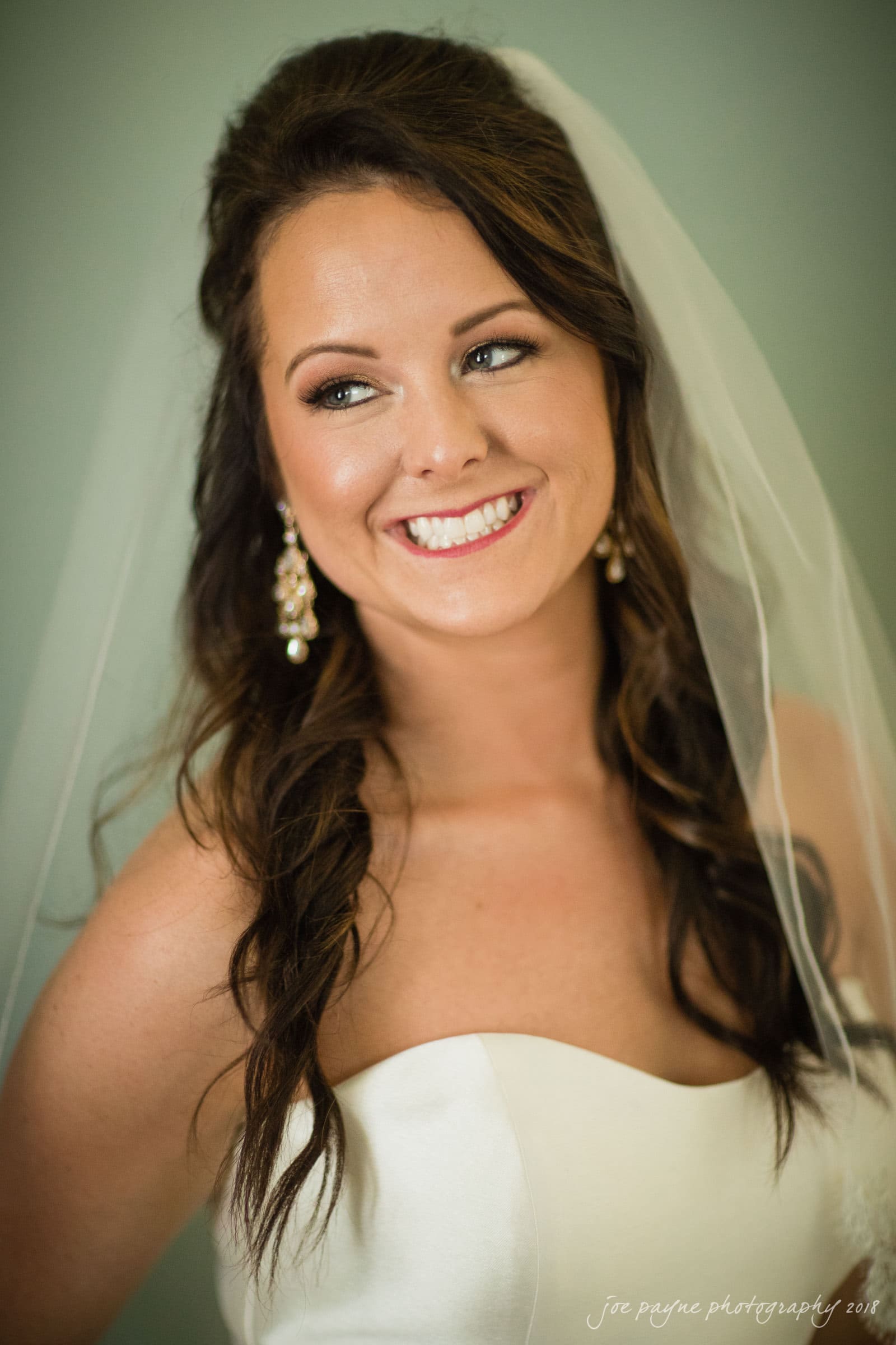may-lew farm wedding photography - macy & jordan A bride wearing a strapless white wedding dress and veil smiles radiantly. Her long brown hair is styled in loose waves, and she wears dangling pearl earrings. The background is softly blurred.