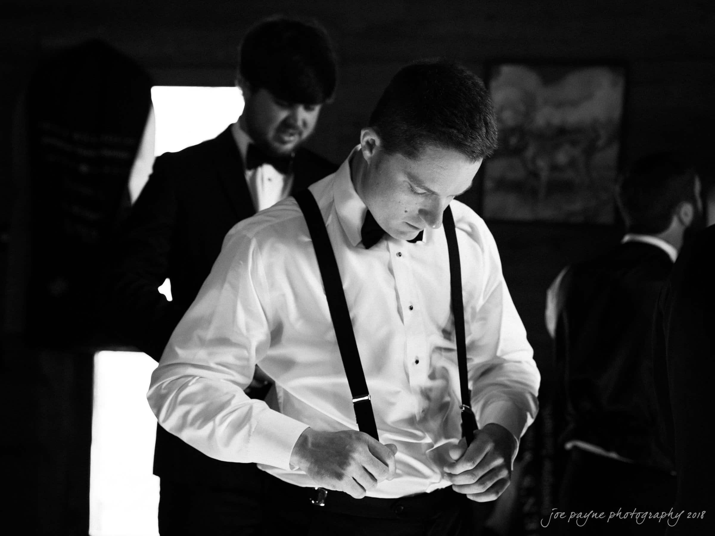 may-lew farm wedding photography - macy & jordan A black and white photo of a man in a white shirt and suspenders adjusting his attire. Another man in the background is wearing a suit, and part of a third person is visible. The setting appears to be indoors.