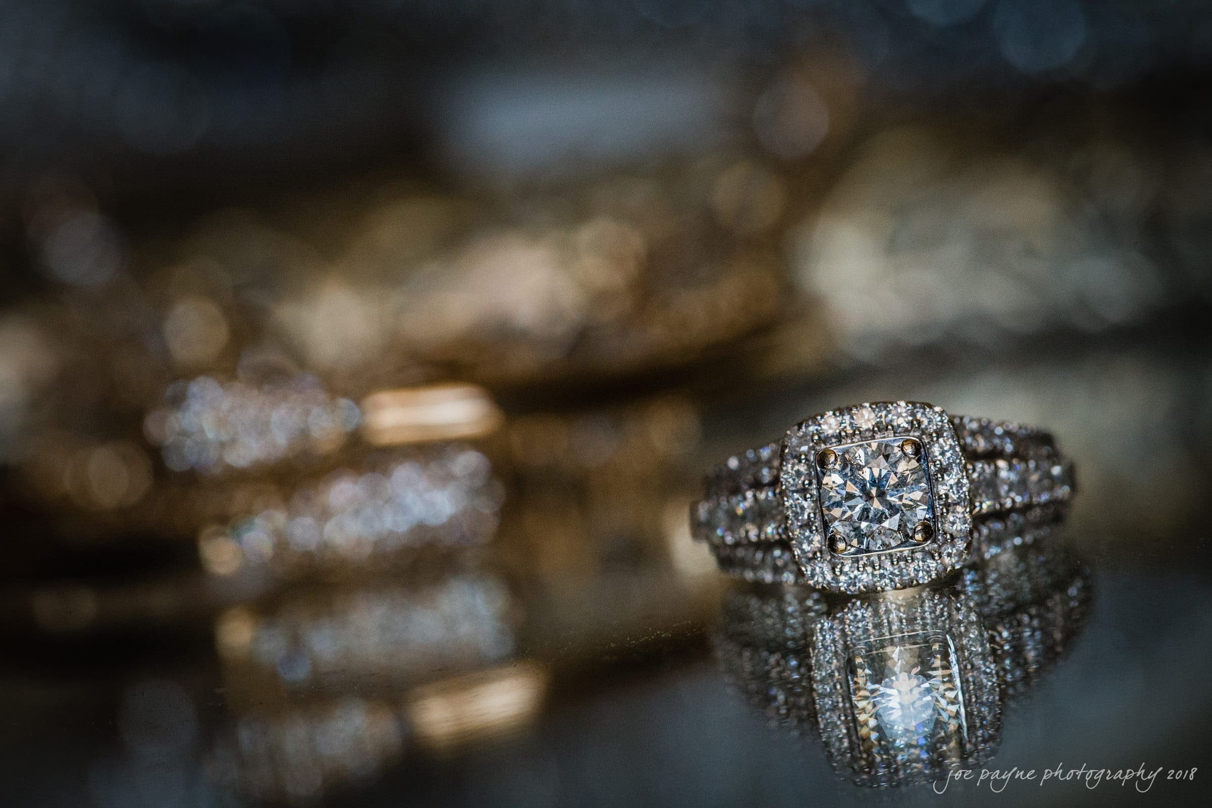 may-lew farm wedding photography - macy & jordan A close-up of a diamond engagement ring with a square halo setting and a double band, reflecting on a shiny surface. The background is blurred, enhancing the focus on the sparkling ring.