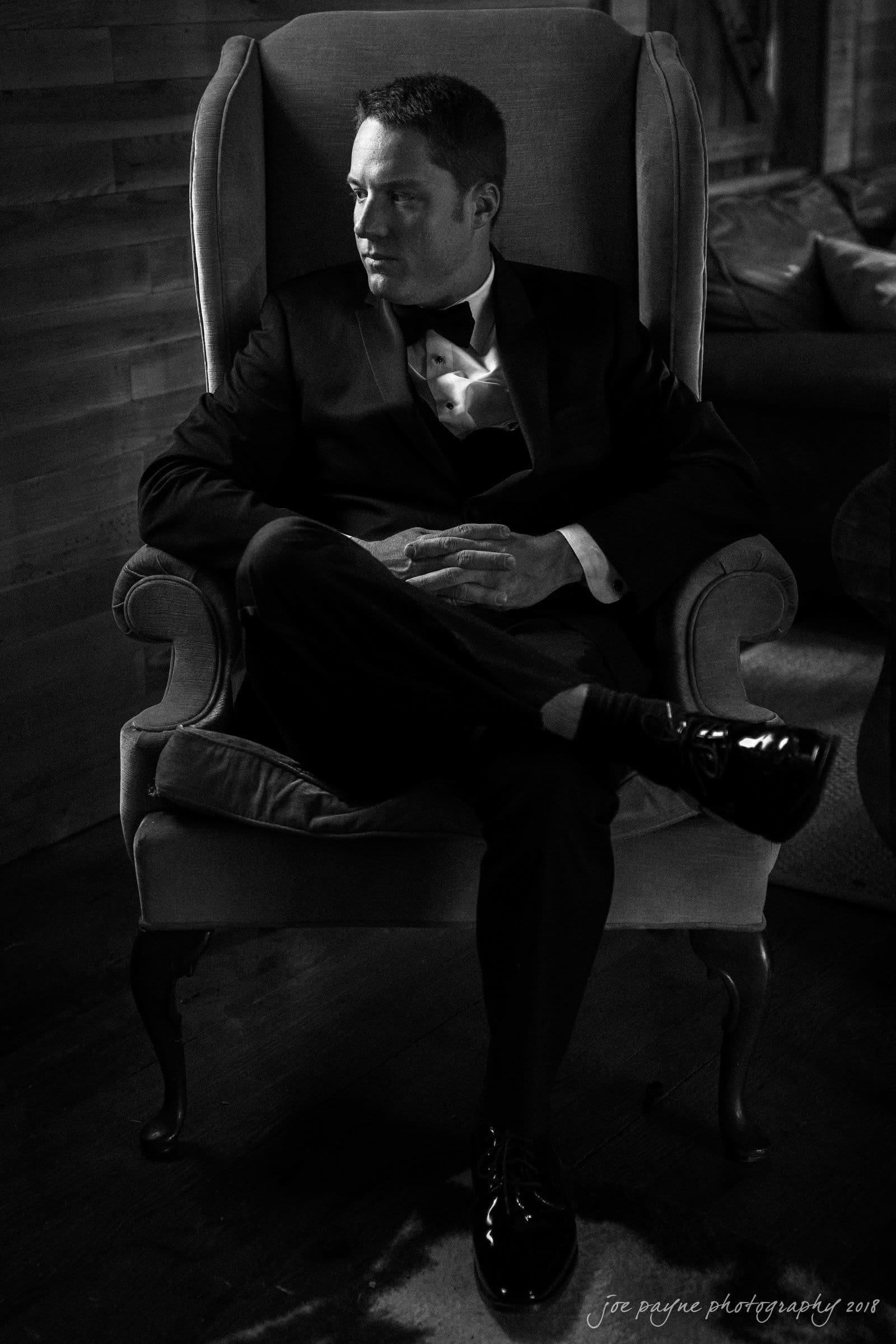 may-lew farm wedding photography - macy & jordan A man in a suit sits in a high-backed armchair, looking to the side. The lighting casts dramatic shadows, emphasizing the elegant and contemplative pose. The setting appears classic and refined.