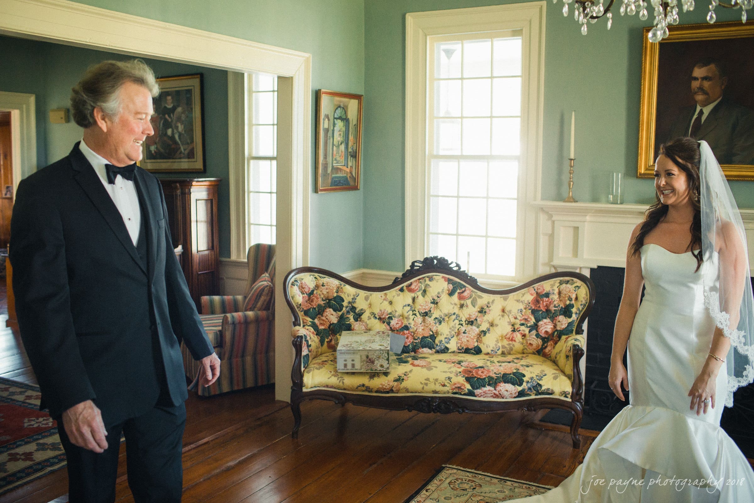 may-lew farm wedding photography - macy & jordan A bride in a white gown and veil smiles at an older man in a tuxedo inside a vintage room with green walls. Elegant furniture, paintings, and a chandelier decorate the space. Natural light filters through large windows.