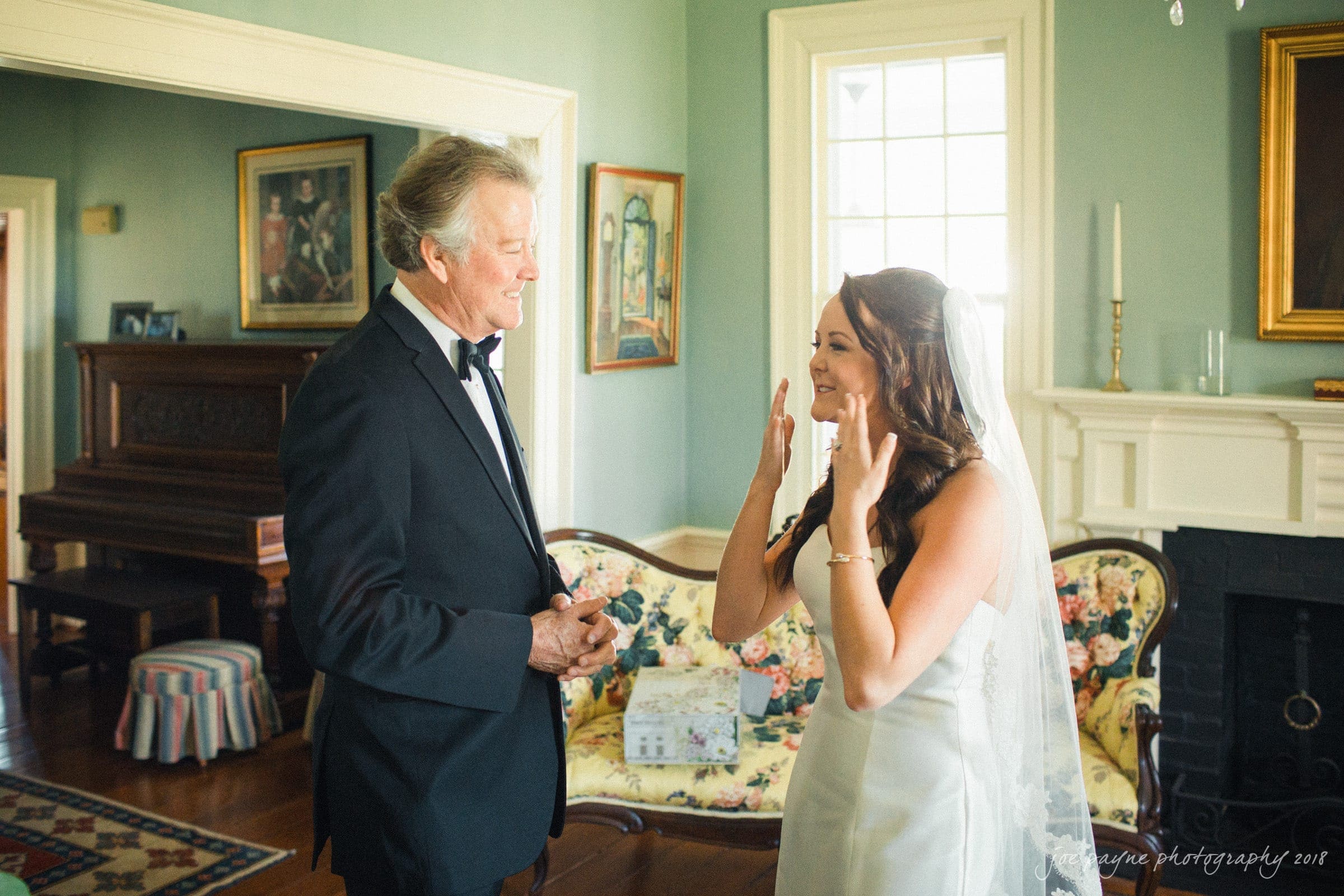 may-lew farm wedding photography - macy & jordan A bride in a white wedding dress and veil is smiling with her hands raised, while an older man in a black suit and bow tie stands beside her, smiling. They are in a living room with elegant decor, including a sofa and a piano.