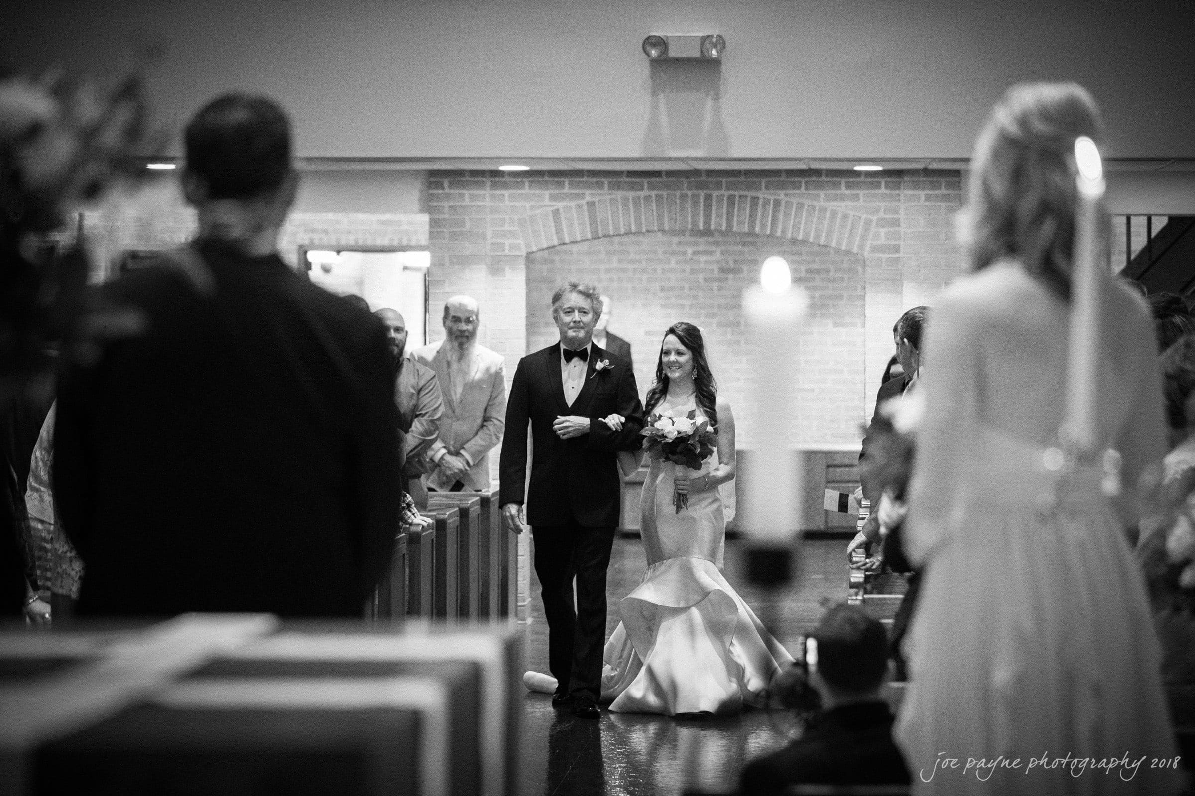 may-lew farm wedding photography - macy & jordan A bride, holding a bouquet, walks down the aisle of a church with an older man in a tuxedo, possibly her father. The aisle is lined with guests seated on either side, and the scene has a black-and-white, formal setting.