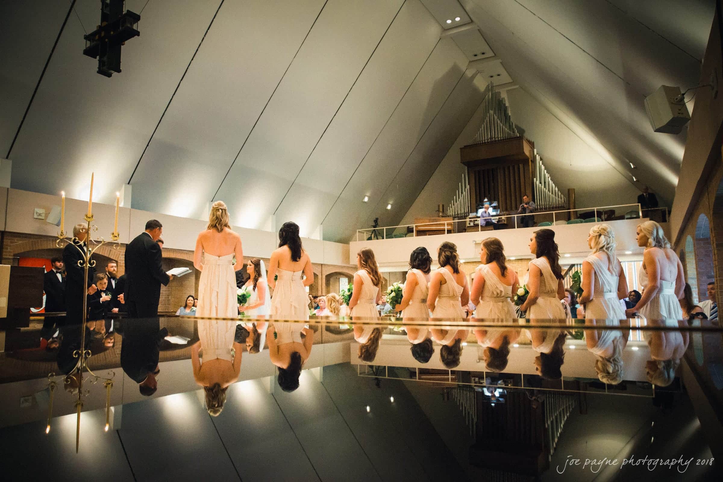may-lew farm wedding photography - macy & jordan A wedding ceremony in a church with a high, sloped ceiling. Bridesmaids in light dresses stand lined up, facing the officiant and groom. Reflections are visible on a polished surface. Candles and an organ are in the background.