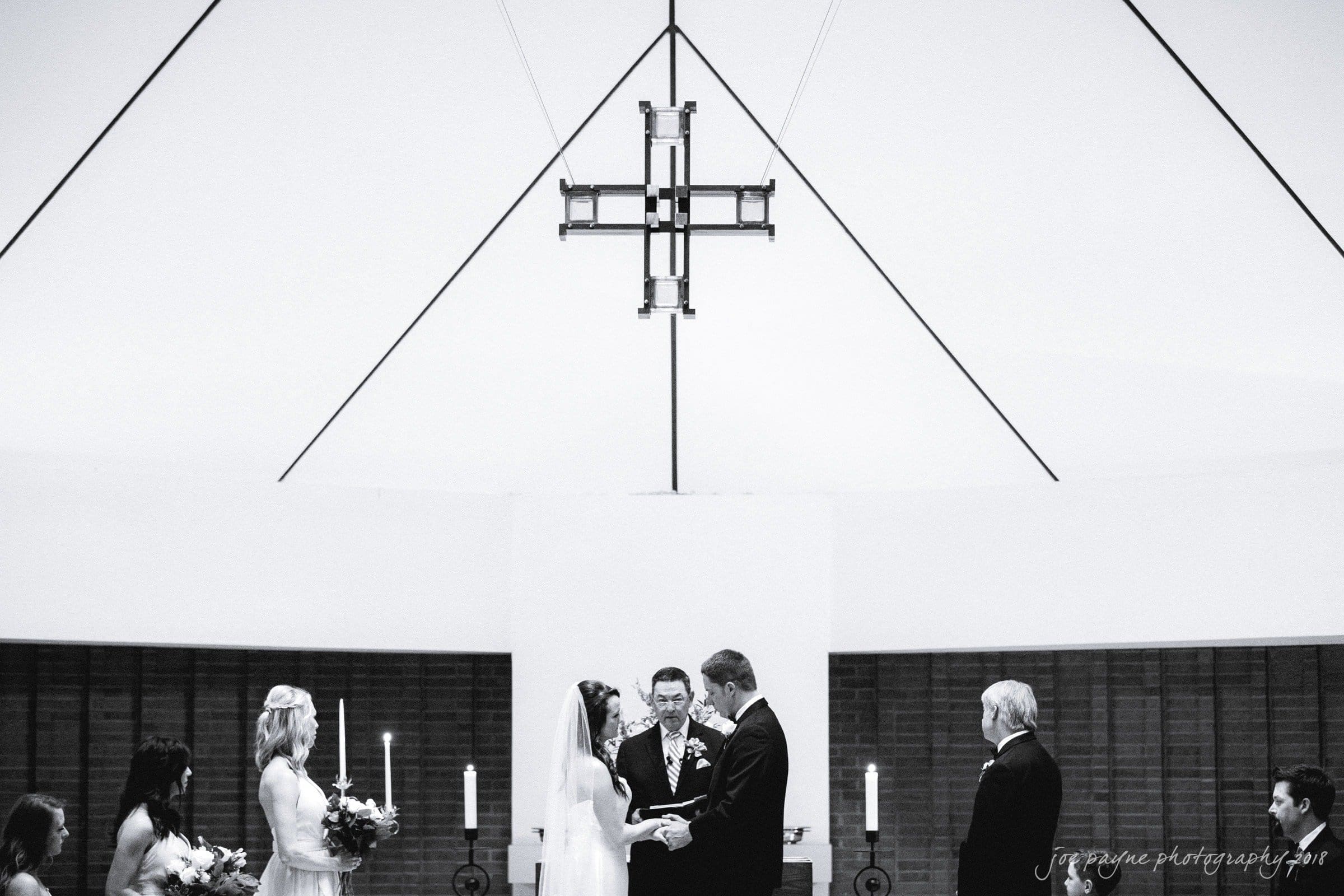 may-lew farm wedding photography - macy & jordan A bride and groom stand facing each other at an altar inside a church, exchanging vows. Bridesmaids and groomsmen are on either side. A large cross hangs above them. Candles are lit in the background.