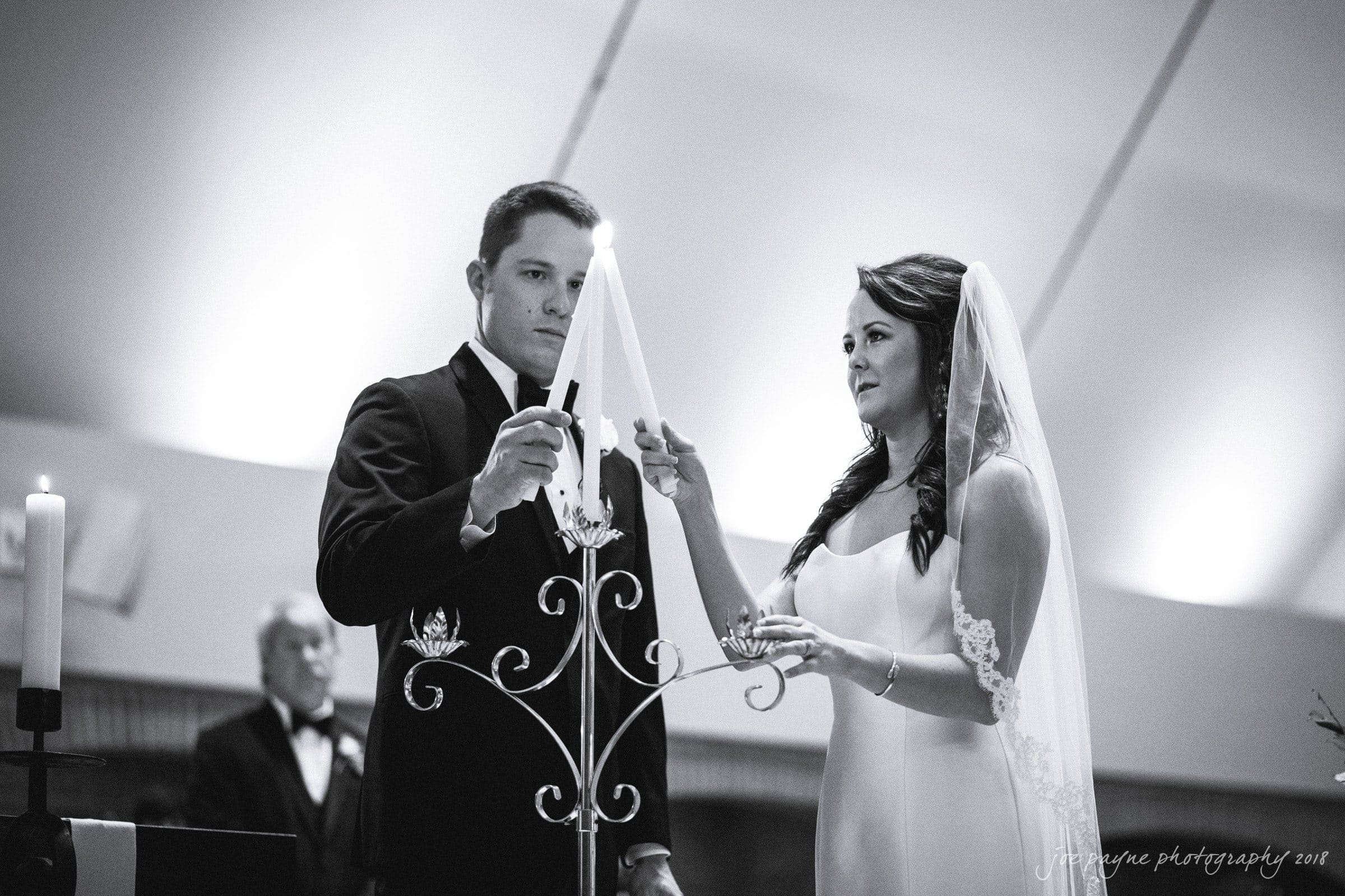 may-lew farm wedding photography - macy & jordan A bride and groom light a candle together during a wedding ceremony. They stand next to a decorative candle holder, with the groom in a tuxedo and the bride in a wedding dress and veil. The setting is elegantly lit.