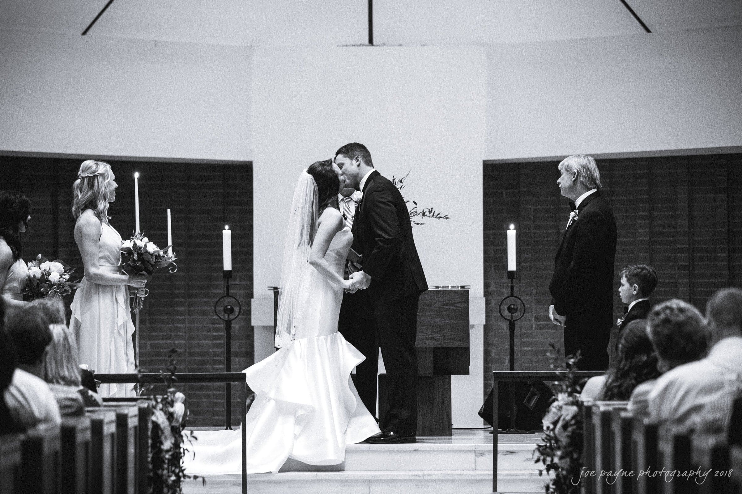 may-lew farm wedding photography - macy & jordan A black and white photo of a wedding ceremony. The bride and groom kiss at the altar, surrounded by the bridal party and guests. Candles and floral arrangements adorn the scene, while the wedding party stands nearby.