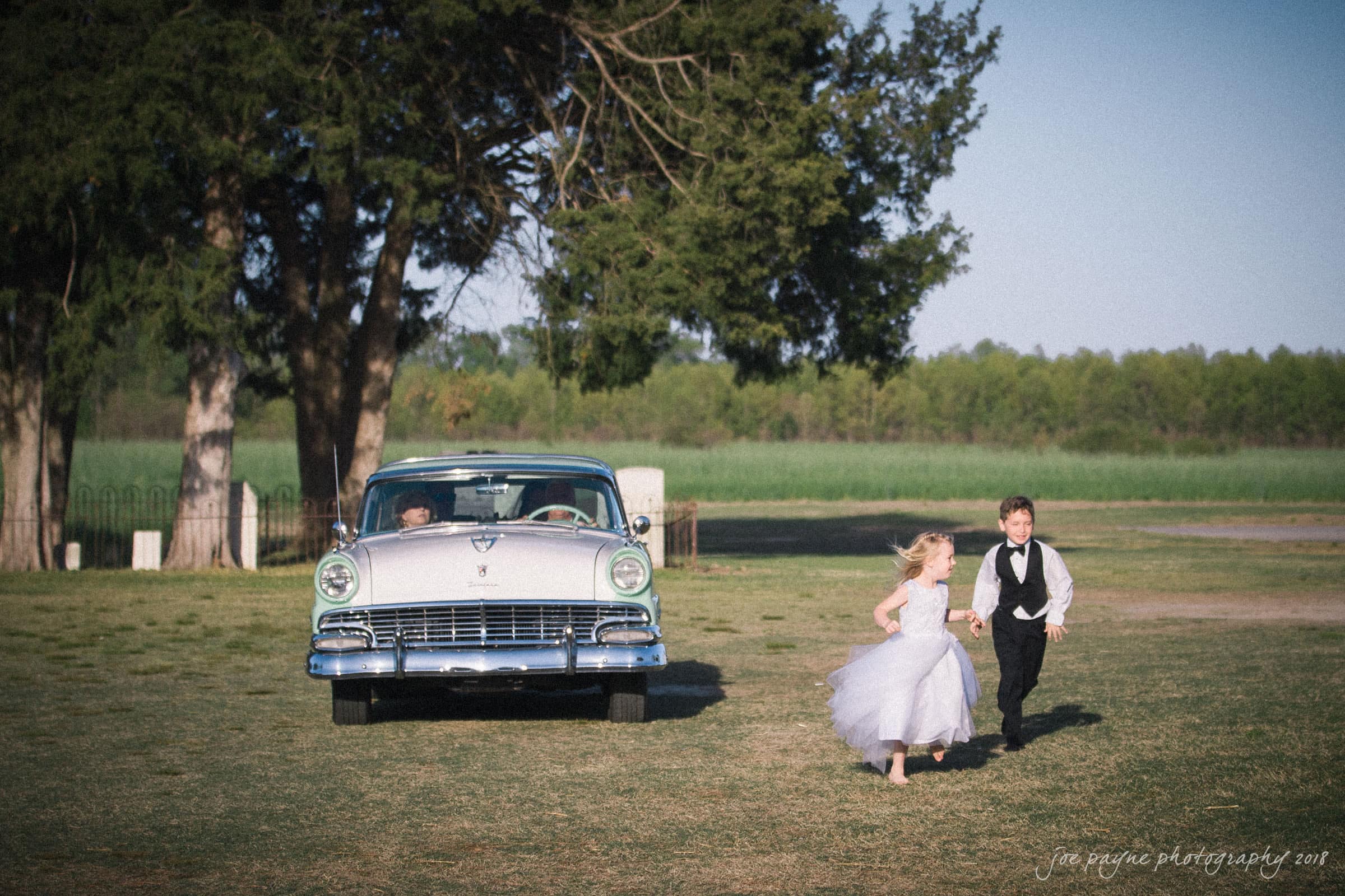 may-lew farm wedding photography - macy & jordan Two children, a boy and a girl, are happily running on grass near a vintage car parked under a tree. The girl is wearing a white dress, and the boy is in formal attire. The background features a lush green field and a clear blue sky.