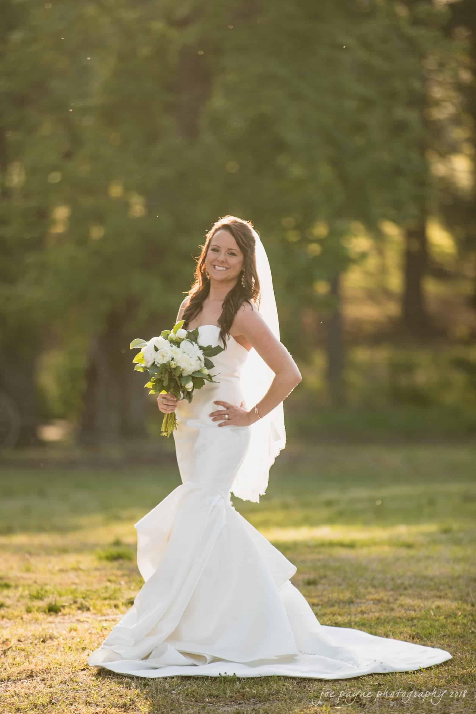 may-lew farm wedding photography - macy & jordan A bride in a flowing white gown and veil stands outdoors on a sunlit lawn, smiling and holding a bouquet of white flowers. The background features lush green trees and soft, warm sunlight.