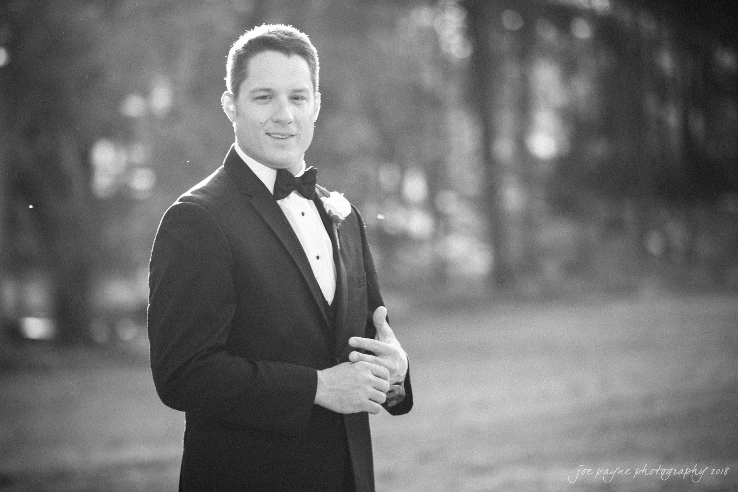 may-lew farm wedding photography - macy & jordan A man in a tuxedo stands outdoors on a sunny day. He adjusts his jacket and looks to the side with a slight smile. The background is softly blurred, highlighting the formal attire and serene setting.
