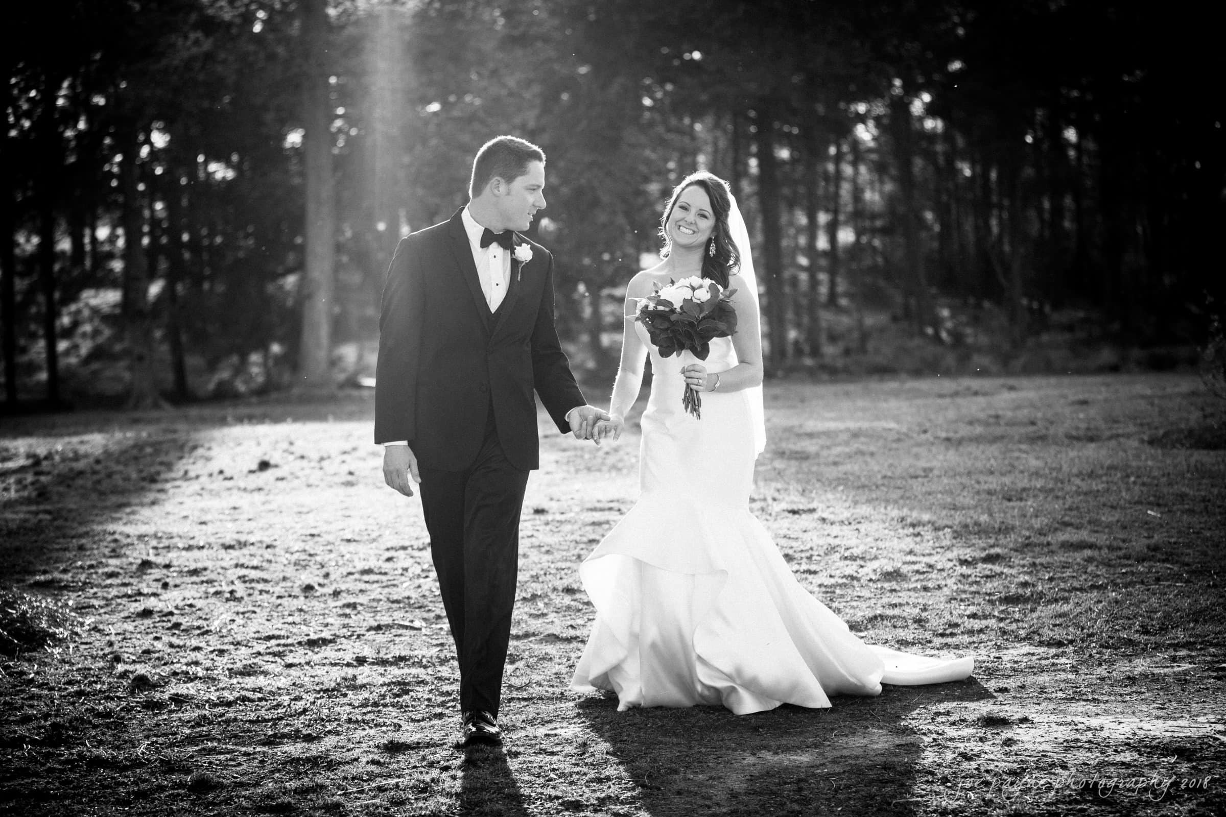 may-lew farm wedding photography - macy & jordan A bride and groom walk hand in hand through a sunlit wooded area. The bride, in a flowing gown, holds a bouquet and smiles. The groom wears a tuxedo, looking at her affectionately. Sunlight filters through the trees, creating a romantic atmosphere.