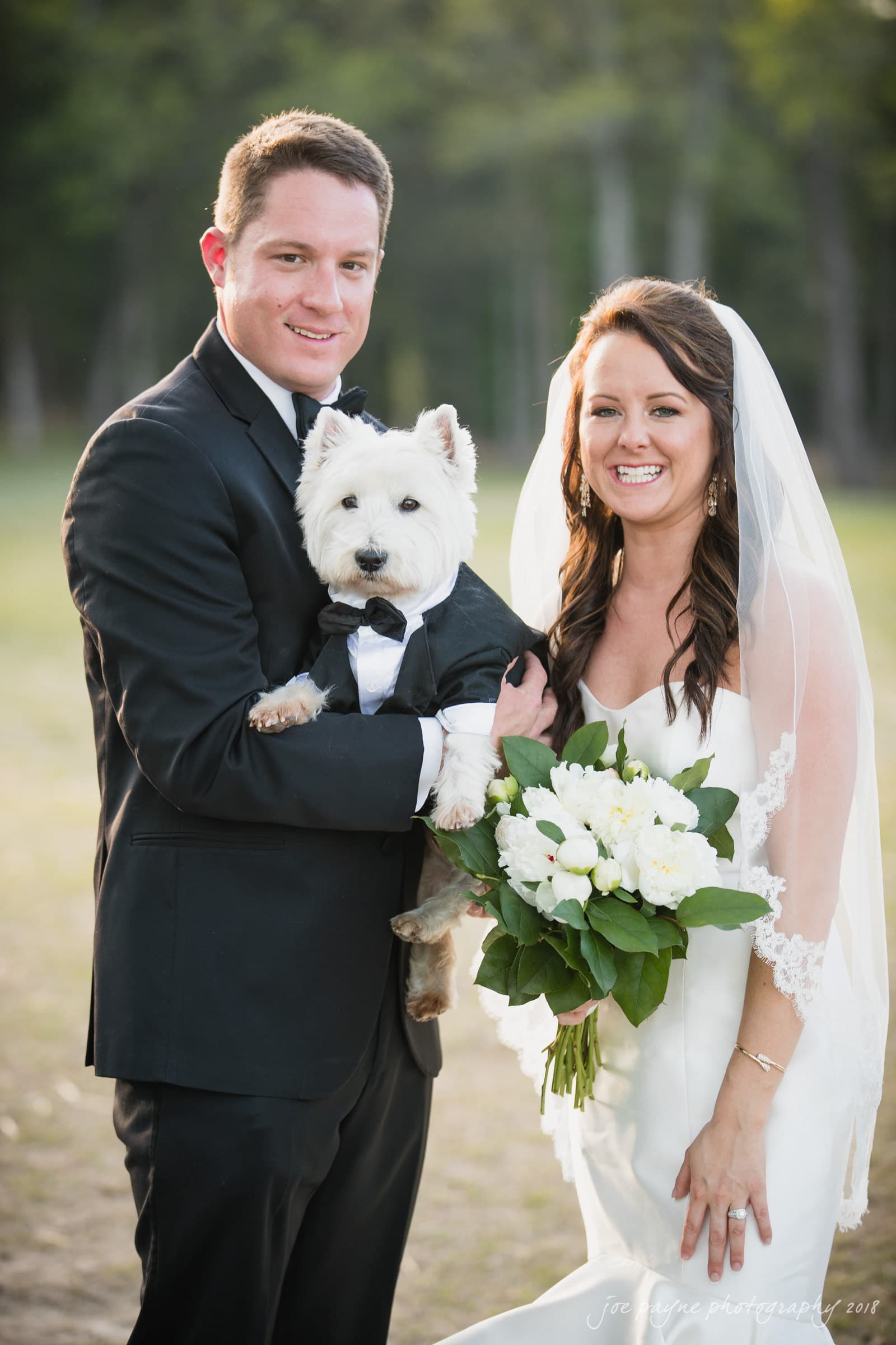 may-lew farm wedding photography - macy & jordan A bride and groom smile while holding a small white dog dressed in a tuxedo. The bride is in a white gown with a veil, holding white flowers. The groom is in a black suit. They are standing outdoors on a sunny day.