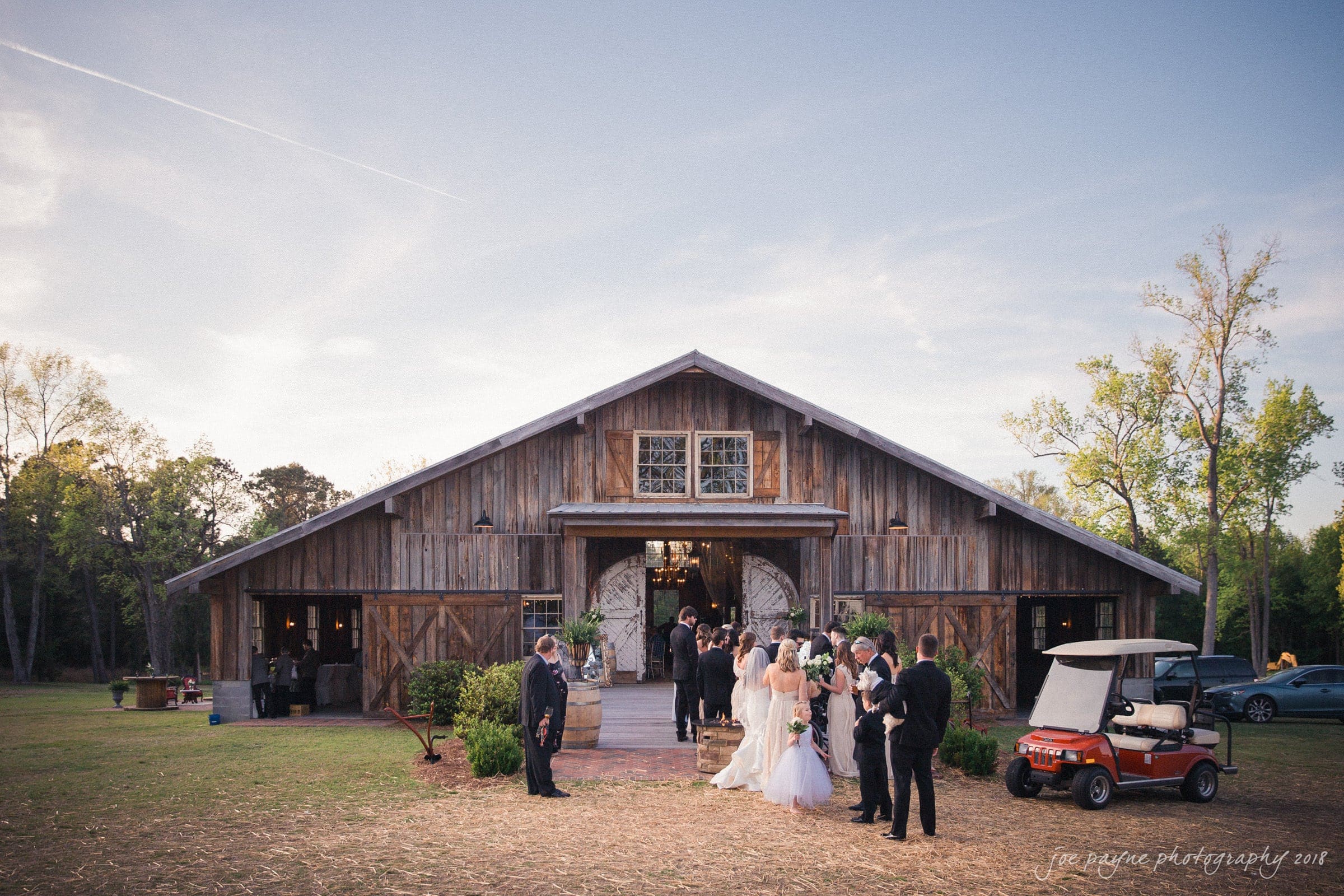 may-lew farm wedding photography - macy & jordan A rustic wedding scene in front of a wooden barn with a group of people dressed in formal attire, including a bride and groom. Trees surround the area, and a golf cart is parked nearby under a clear sky.