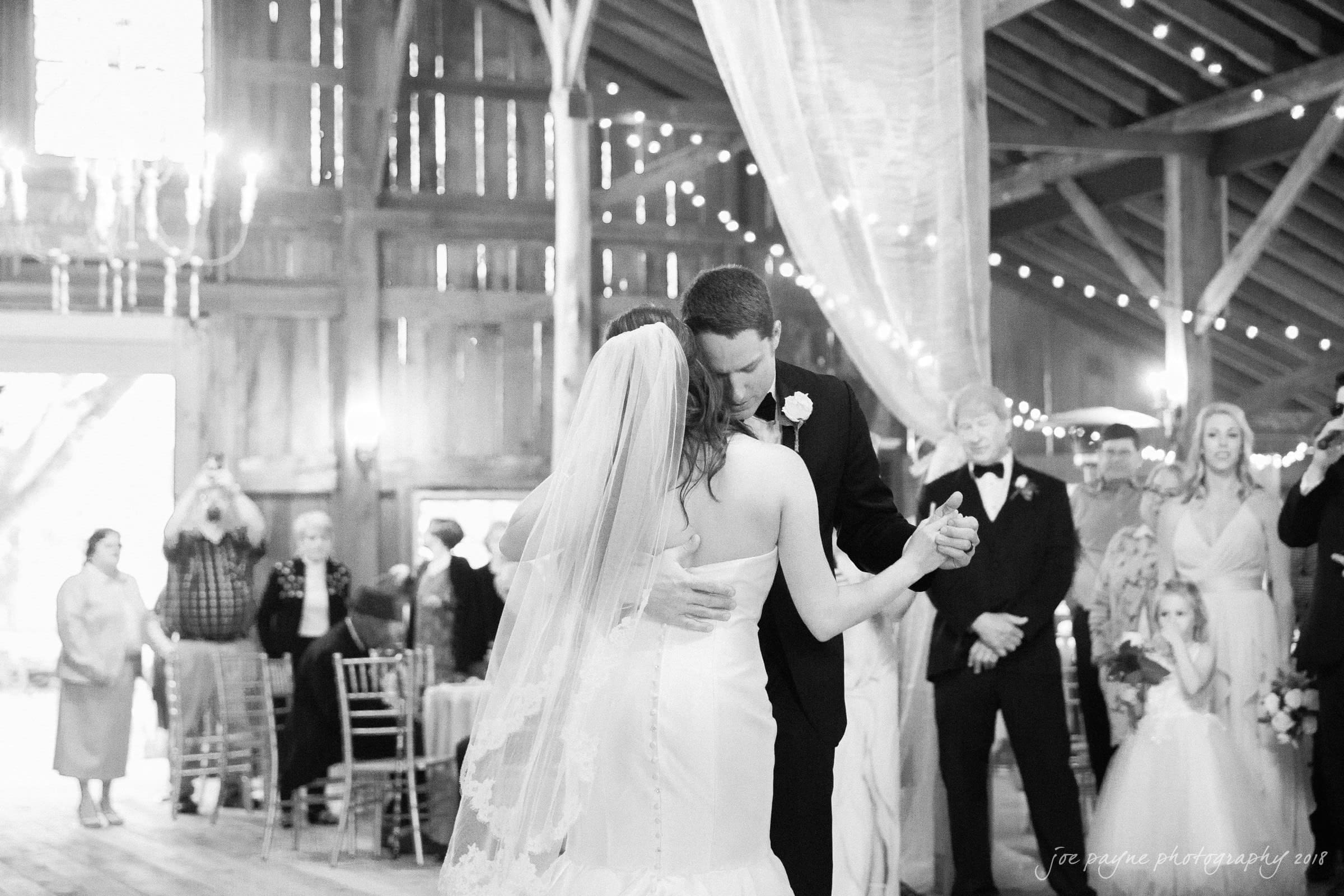 may-lew farm wedding photography - macy & jordan A bride and groom share their first dance in a rustic barn setting. String lights and a chandelier add a warm glow, while guests watch in the background. The bride wears a veil and strapless gown, and the groom is in a suit.