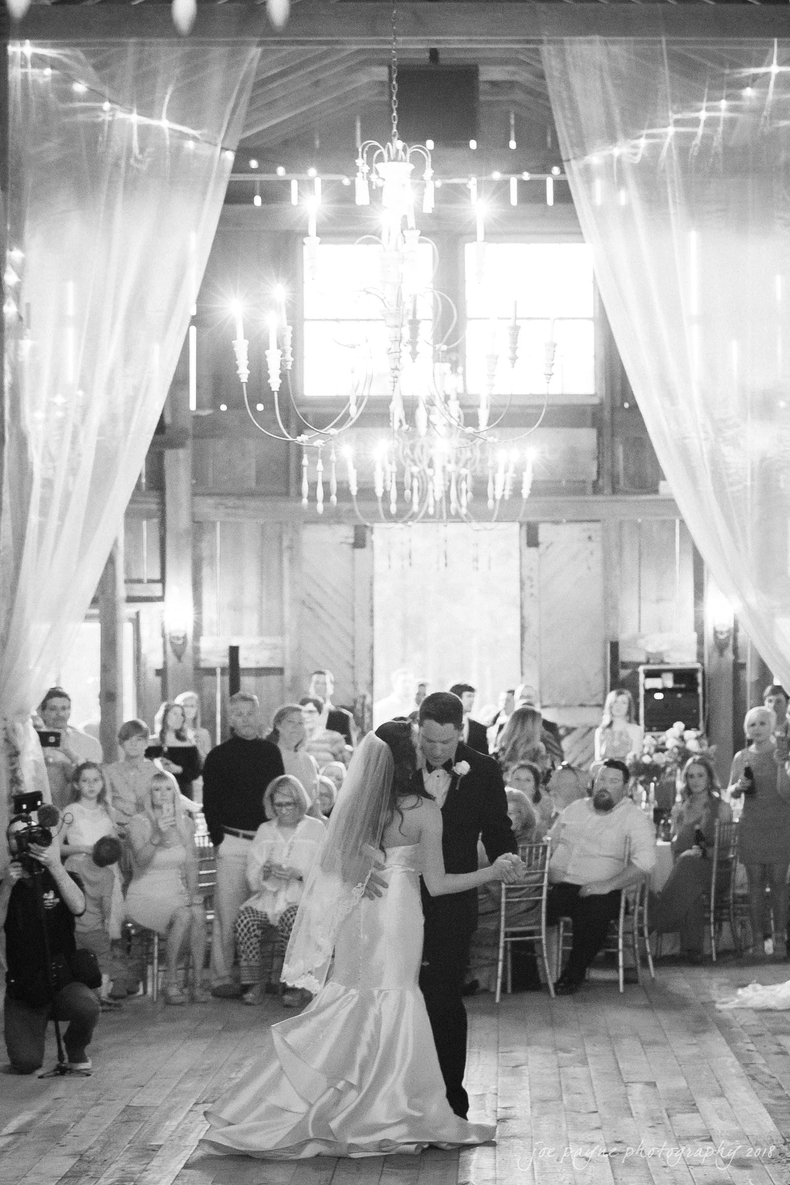may-lew farm wedding photography - macy & jordan A bride and groom share their first dance in a rustic venue with wooden floors and draped curtains. A chandelier hangs overhead, and guests are seated around them, watching the couple. The setting has a romantic and intimate atmosphere.