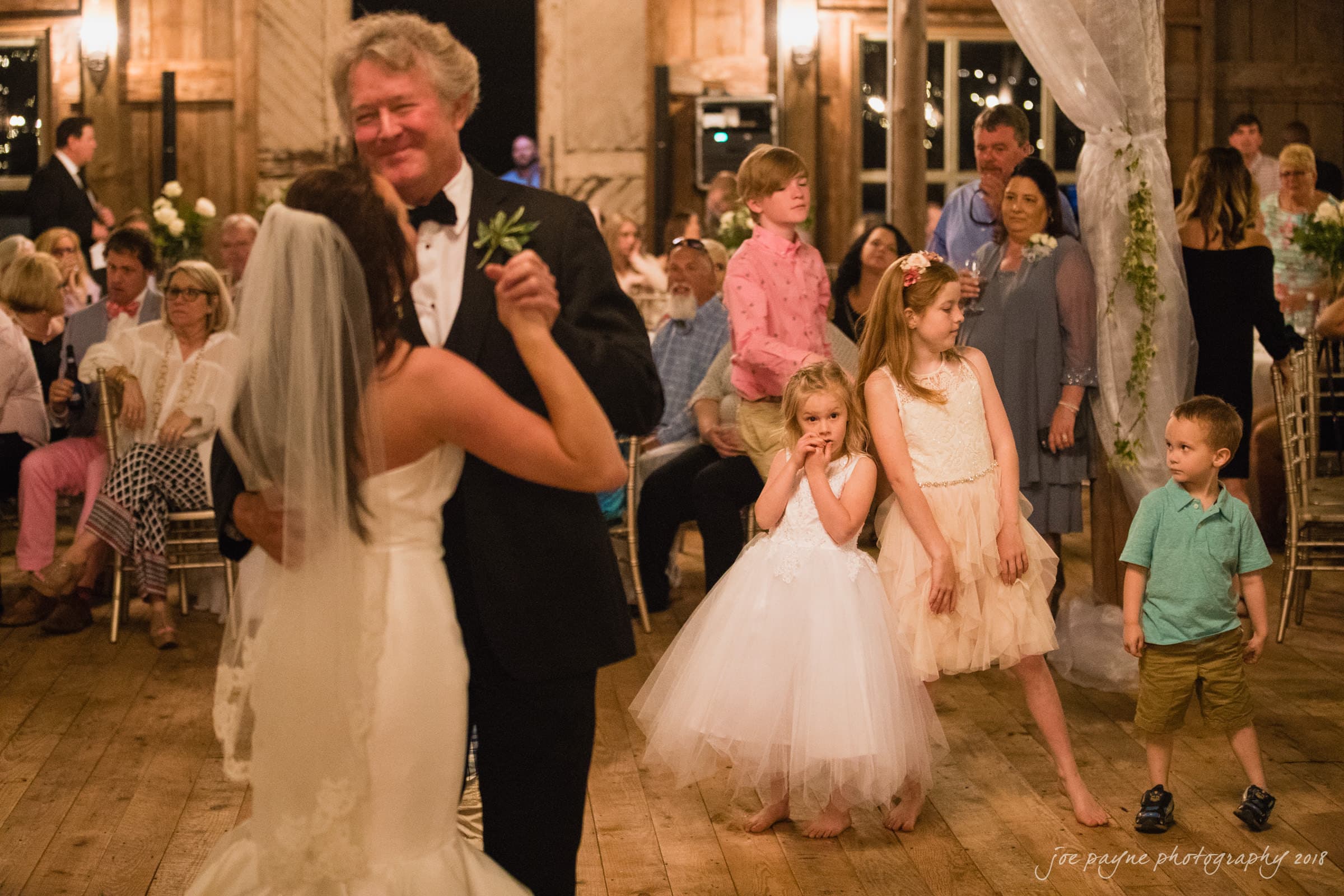 may-lew farm wedding photography - macy & jordan A bride and an older man dance at a rustic indoor wedding reception. Guests watch, including two young girls in white dresses and a boy in the background. The atmosphere is warm and festive.