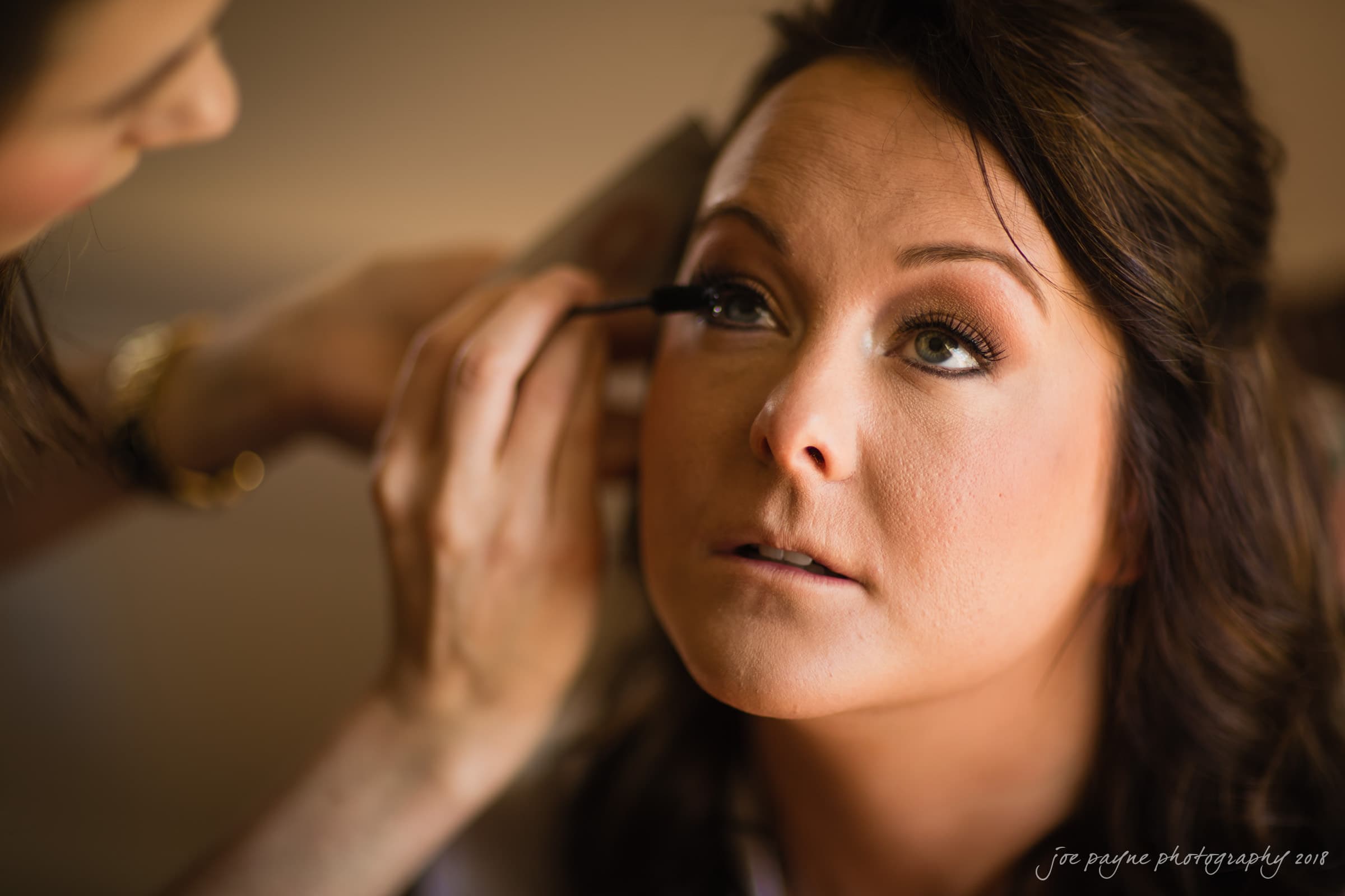 may-lew farm wedding photography - macy & jordan A woman with dark brown hair gets her makeup done, with another person applying mascara to her eyelashes. She gazes upward with a calm expression, and the background is softly blurred.