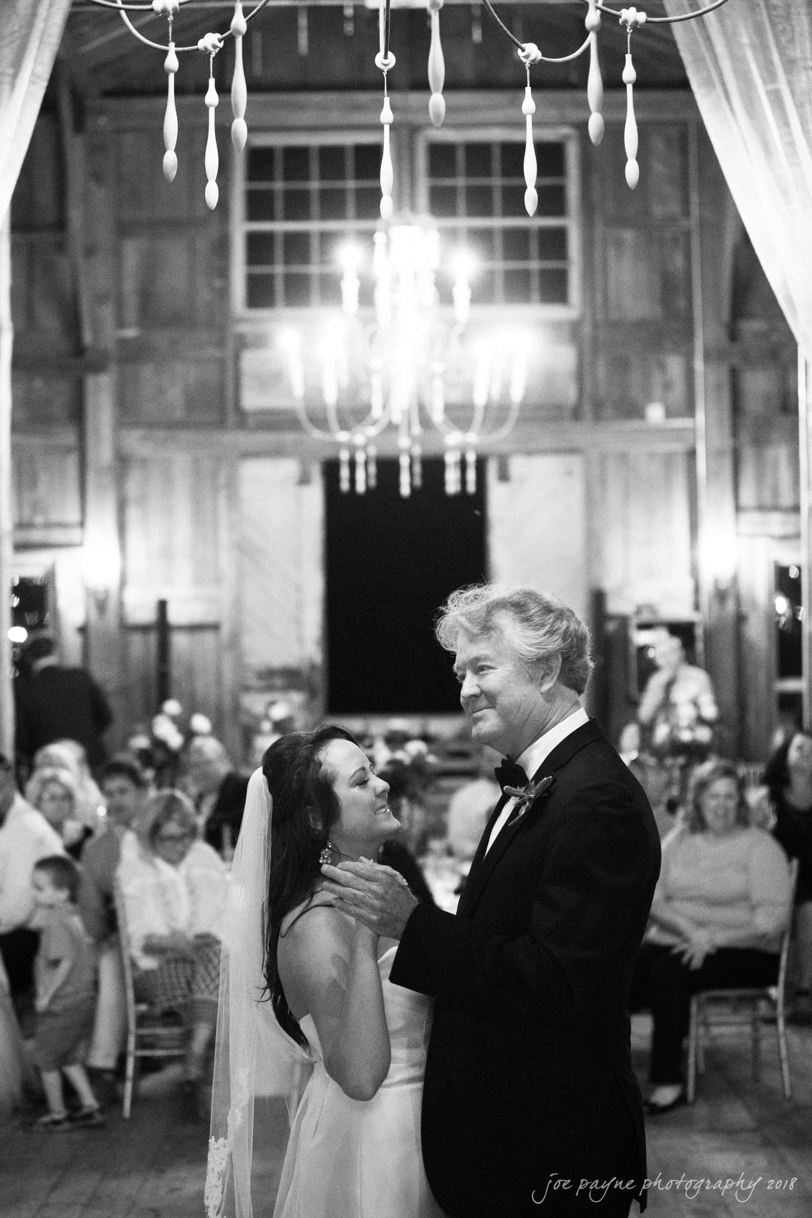 may-lew farm wedding photography - macy & jordan A bride and groom share a first dance in a warmly lit, rustic venue. They are surrounded by seated guests. The room features wooden walls and a large chandelier.
