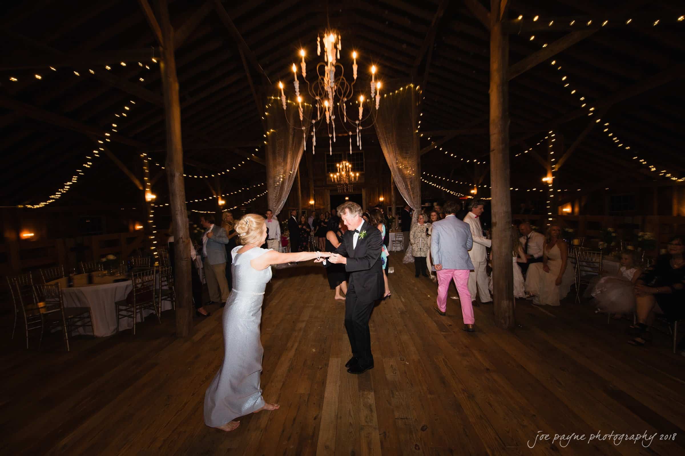 may-lew farm wedding photography - macy & jordan A couple dances on a wooden floor under a string of lights and chandeliers in a barn setting. Guests are seated around the room, watching. The atmosphere is warm and festive.