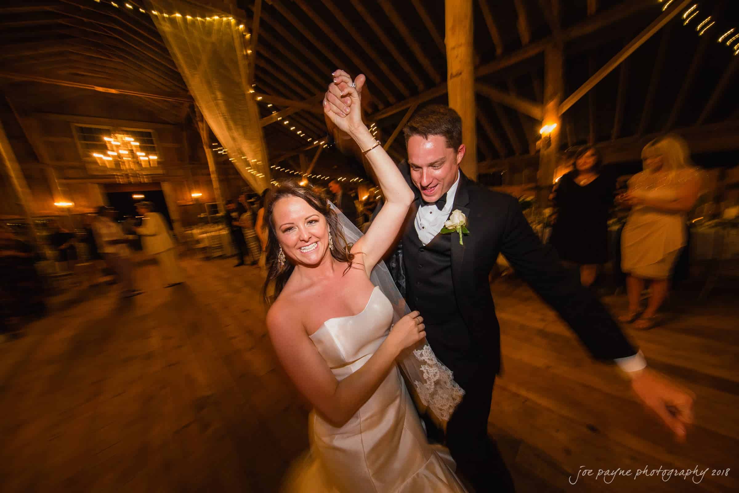may-lew farm wedding photography - macy & jordan A bride and groom dance joyfully in a warmly lit barn, with wooden beams and string lights in the background. The bride wears a strapless white gown, and the groom is in a black tuxedo. They hold hands, spinning with big smiles.