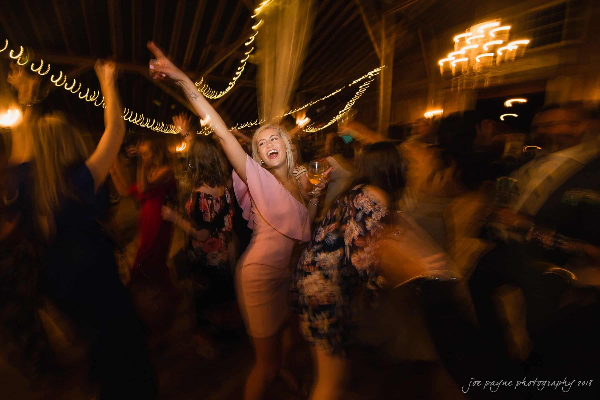 may-lew farm wedding photography - macy & jordan A joyful group of people dancing at a lively party, with one woman in the center wearing a pink dress, holding a drink and raising her arm. The background is blurred with warm, festive lighting and swirling motion.