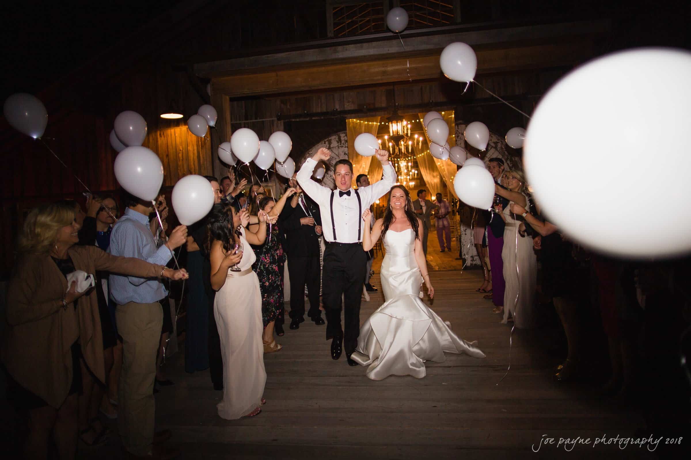 may-lew farm wedding photography - macy & jordan A bride and groom joyfully exit a venue at night, surrounded by cheering guests holding white balloons. The bride is in a white gown, and the groom wears suspenders and a bow tie. The scene is warmly lit with chandeliers in the background.