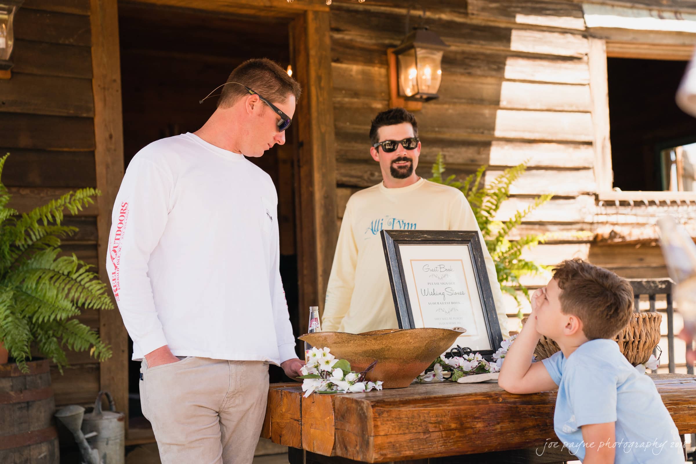 may-lew farm wedding photography - macy & jordan Two men and a boy are gathered around a rustic table outdoors. One man is wearing sunglasses and a white shirt, looking at a framed item on the table. The boy leans on the table, gazing up. A wooden building and ferns are in the background.
