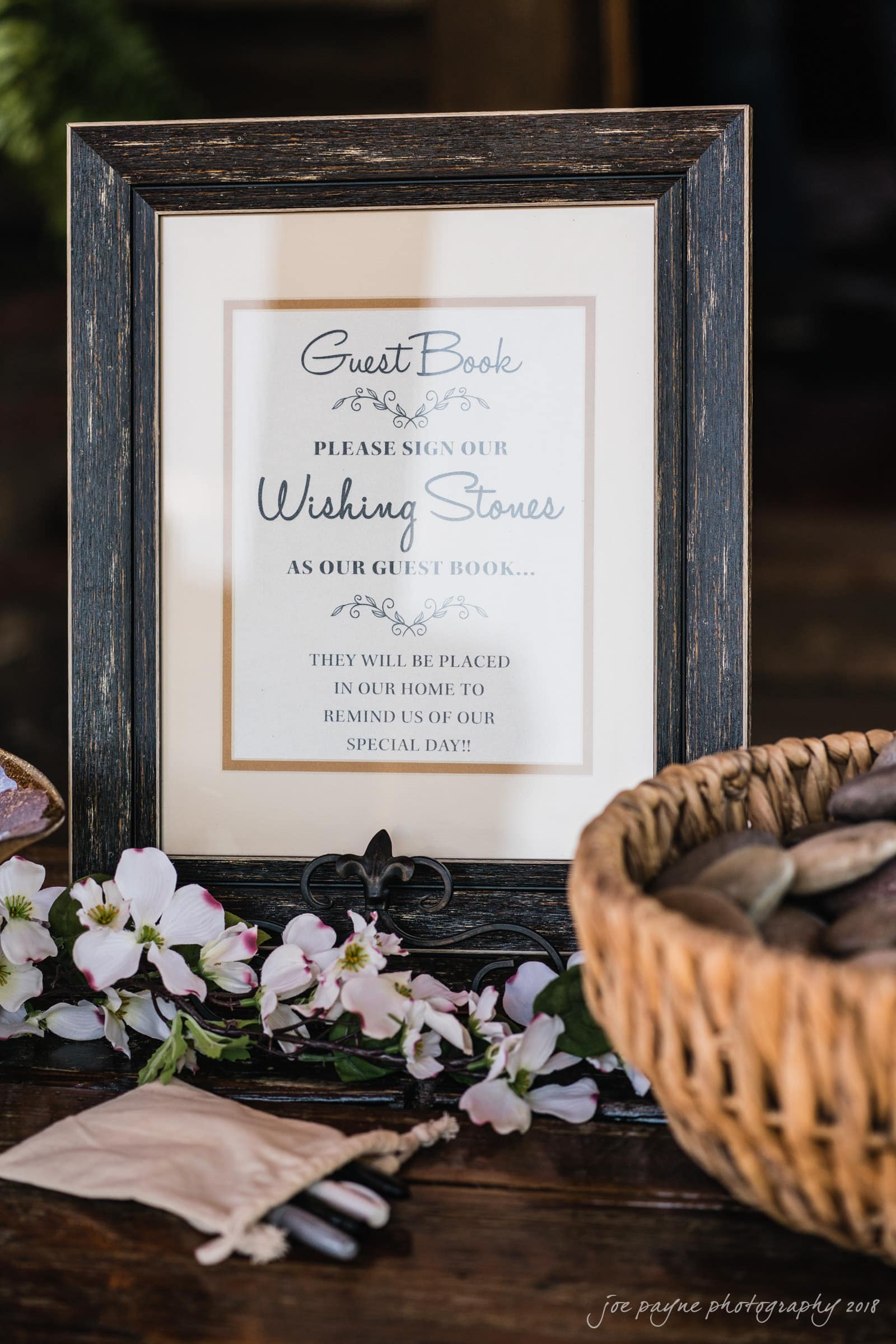 may-lew farm wedding photography - macy & jordan A framed sign invites guests to sign wishing stones as a guest book alternative. The setup includes a basket of stones, a pouch, pens, and decorative flowers. The sign is displayed on a table with a rustic, elegant theme.