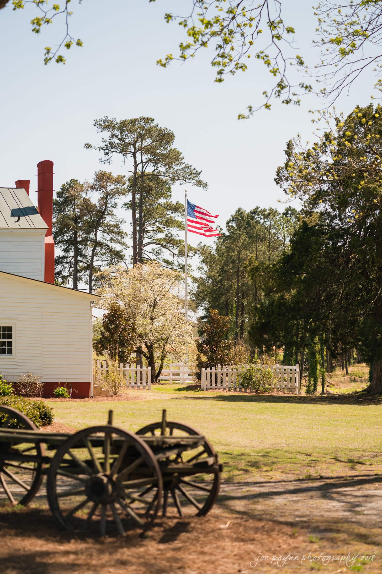 may-lew farm wedding photography - macy & jordan A rustic scene with a wooden wagon wheel in the foreground, an American flag on a flagpole, and a white house with a red roof surrounded by trees and a picket fence in the background.