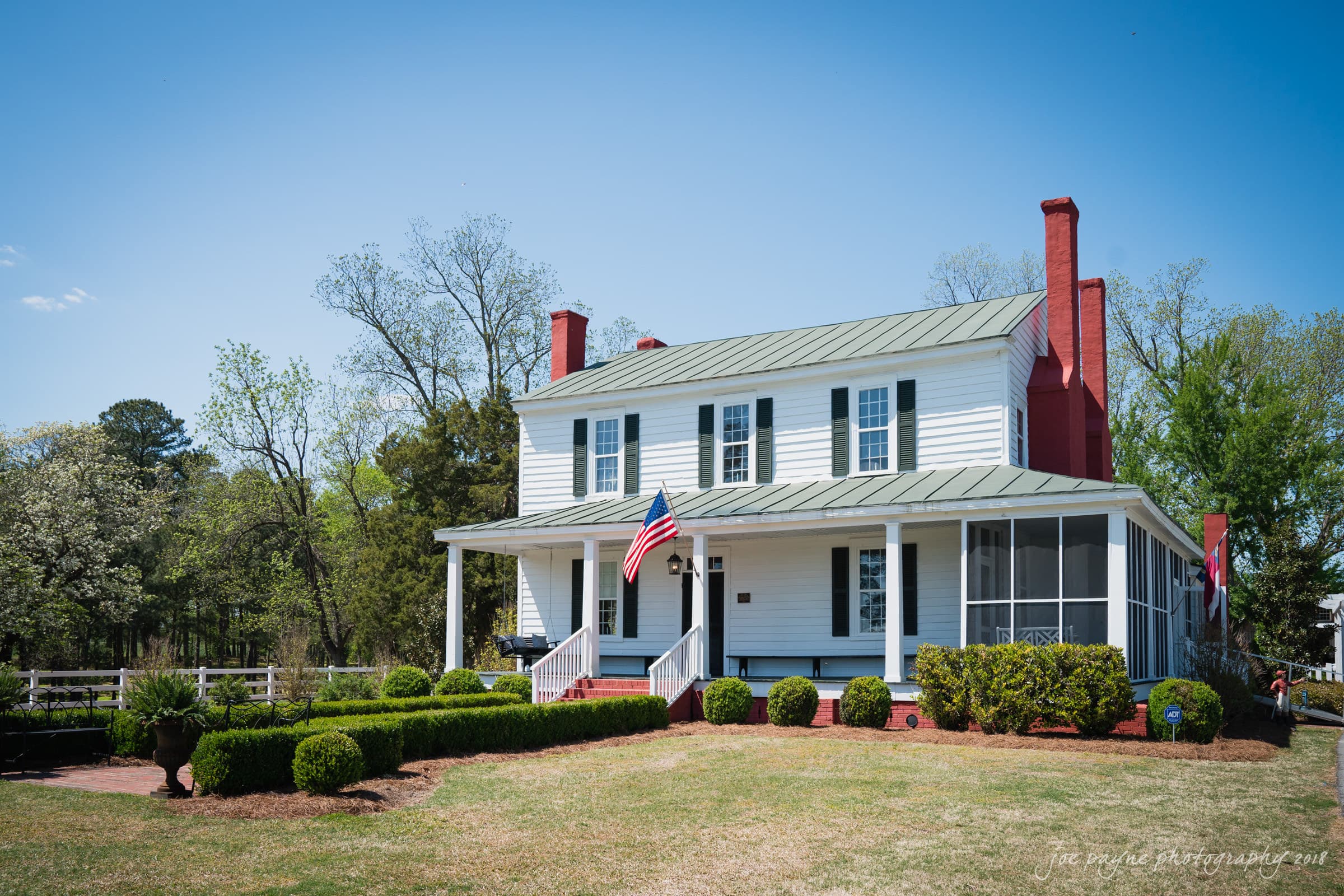 may-lew farm wedding photography - macy & jordan A two-story white house with a green roof and red chimneys, featuring a screened porch and an American flag by the entrance. The yard is landscaped with neatly trimmed bushes, and the background includes tall trees under a clear blue sky.