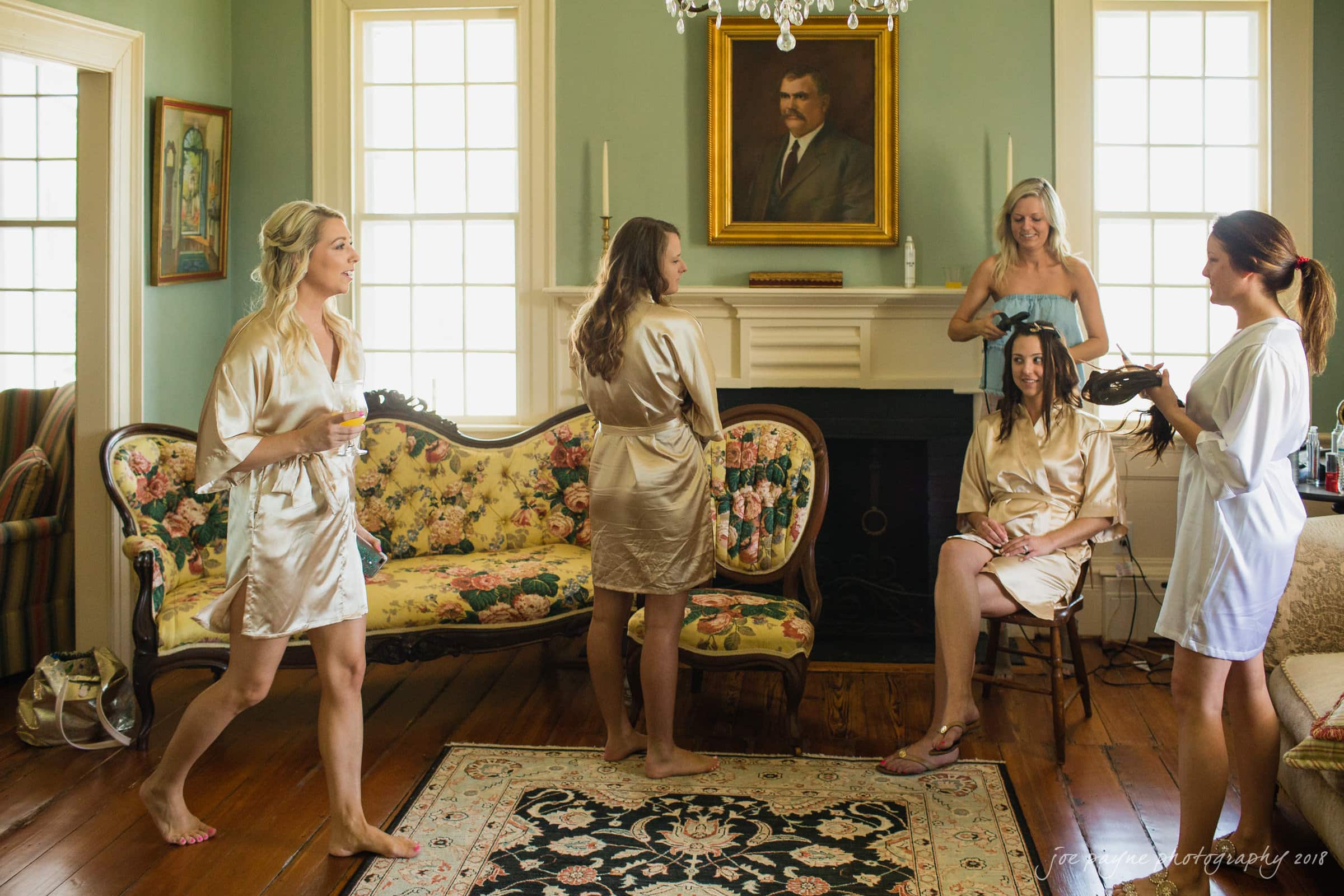 may-lew farm wedding photography - macy & jordan A group of women in satin robes gather in a vintage-style room with wooden floors. One woman sits while having her hair styled. Floral furniture and a large portrait on the wall enhance the settings classic ambiance.