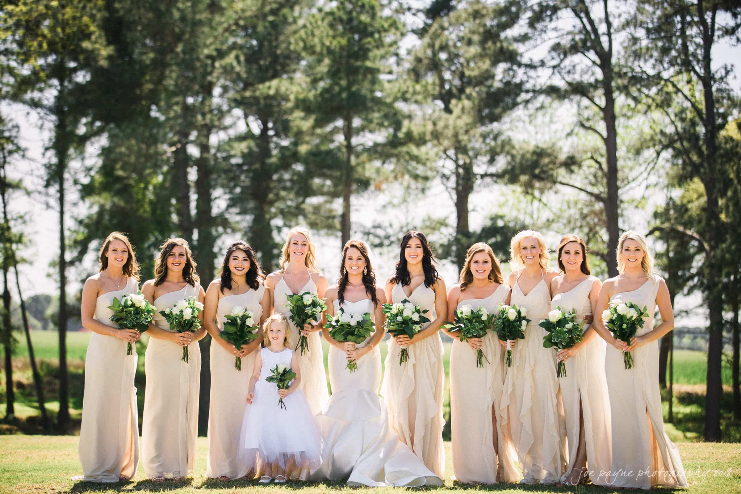 may-lew farm wedding photography - macy & jordan A bridal party poses outdoors in front of trees. Eleven bridesmaids wear matching light-colored dresses and hold white bouquets. A young flower girl in a white dress stands in front, holding a smaller bouquet.