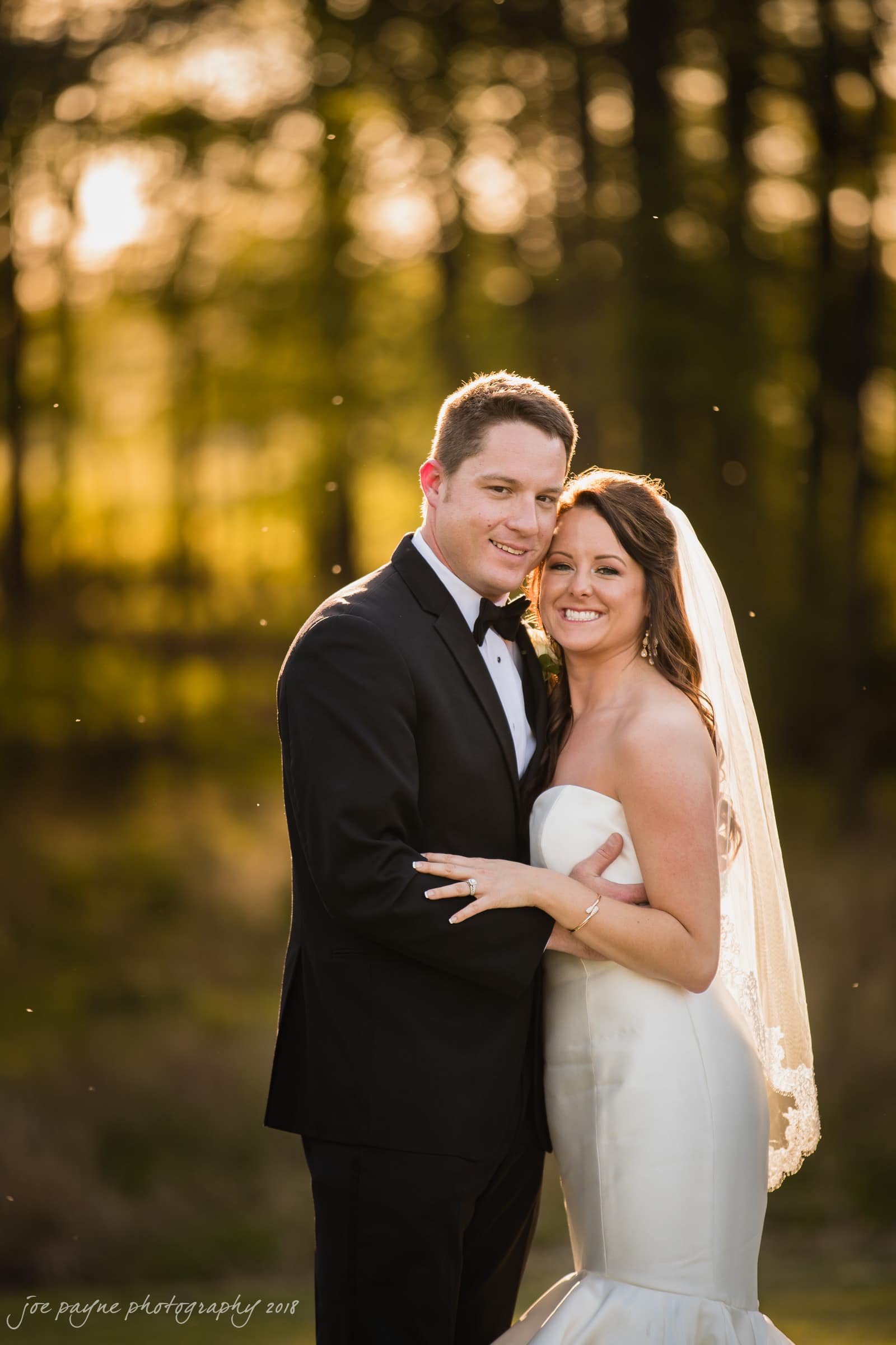 may-lew farm wedding photography - macy & jordan A bride and groom stand closely together outdoors, smiling at the camera. The groom wears a black tuxedo, and the bride is in a white strapless dress with a veil. Sunlight filters through the trees in the background.