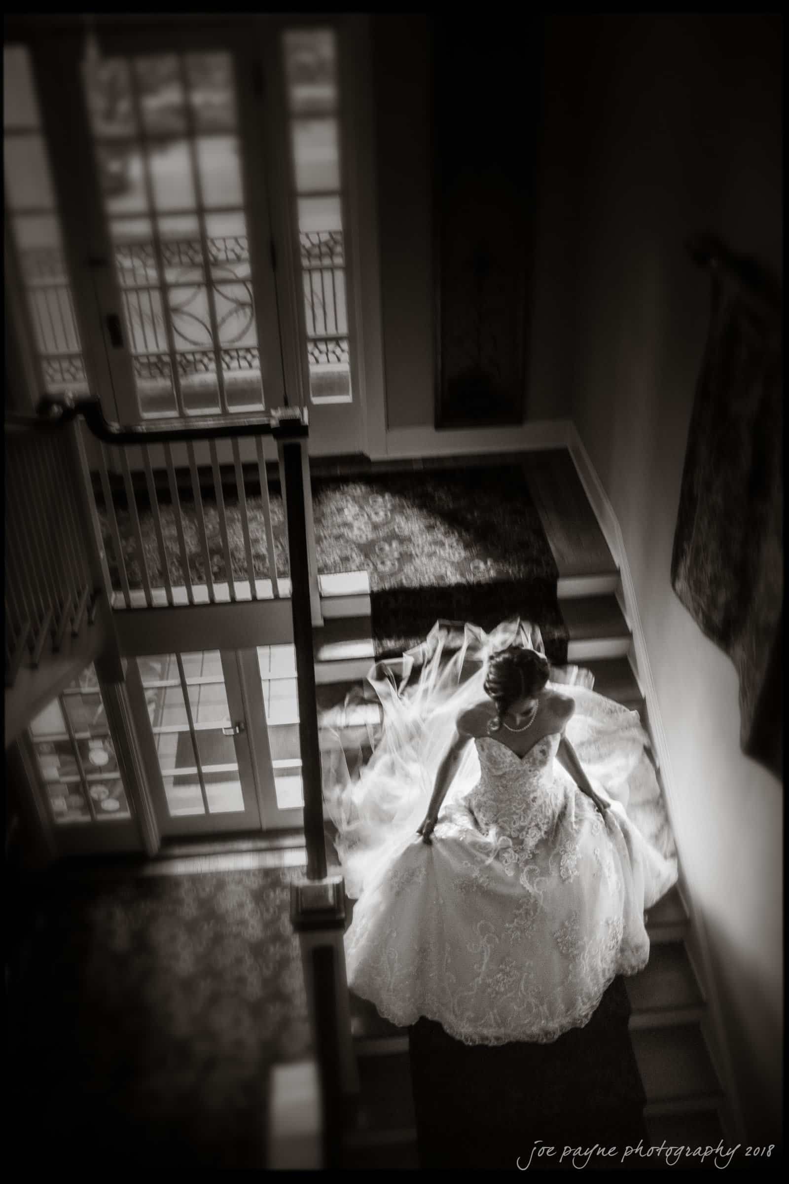 A bride in a white lace gown sits gracefully on a staircase, illuminated by window light from above. She holds her veil, creating an elegant and serene atmosphere.