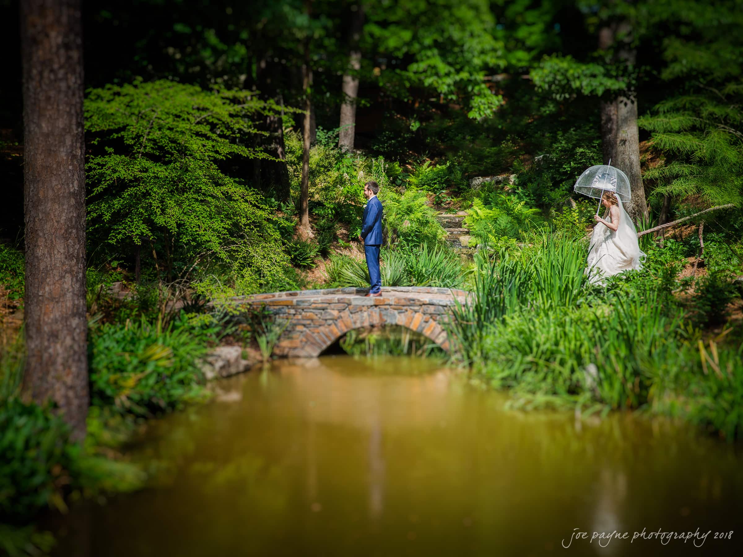 A bride holding an umbrella walks across a stone bridge towards a groom in a blue suit. They are surrounded by lush greenery and a small pond below the bridge.