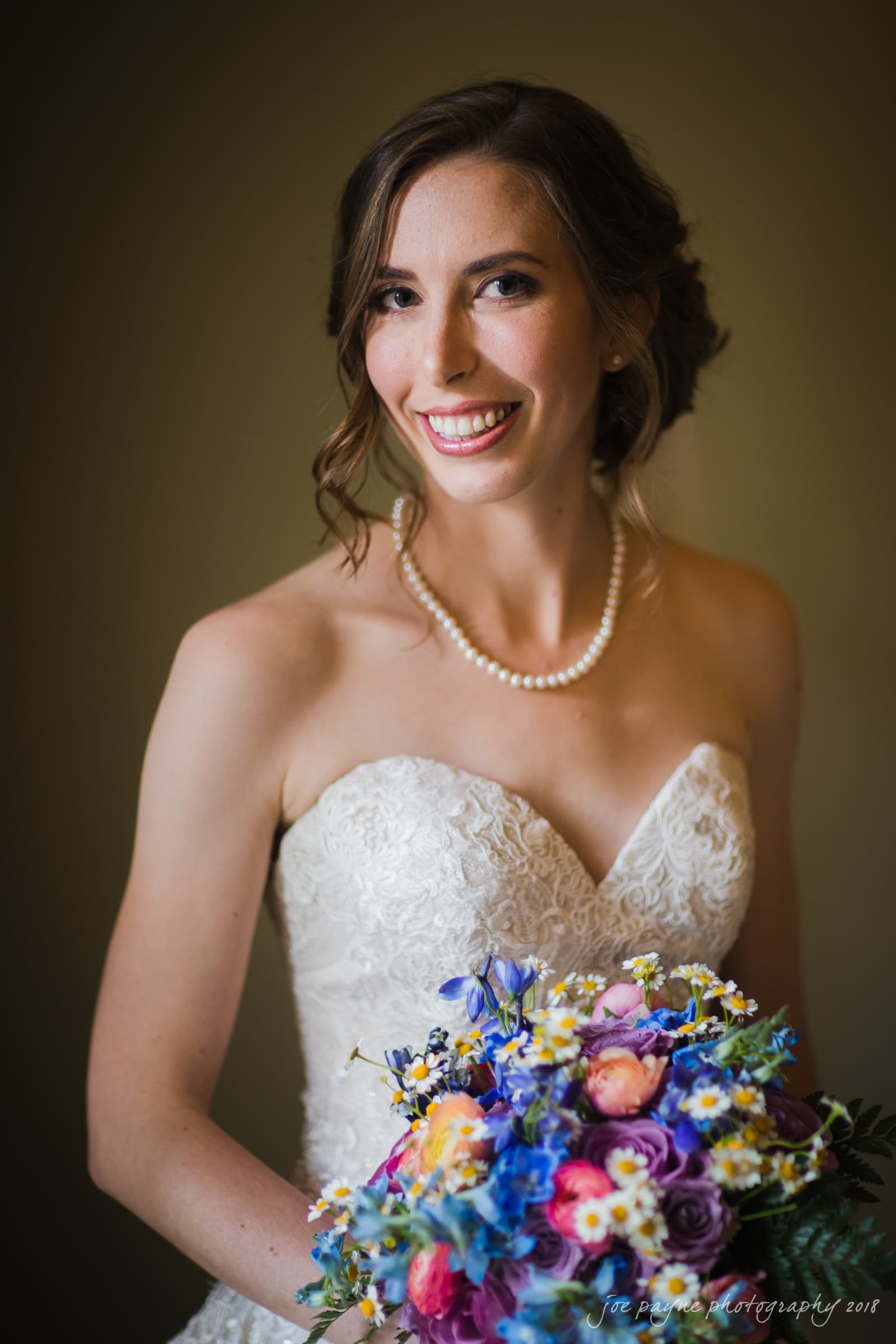 A bride in a white lace strapless gown smiles while holding a colorful bouquet of flowers, including blue, purple, and yellow blooms. She wears a pearl necklace and has her hair styled in soft waves.