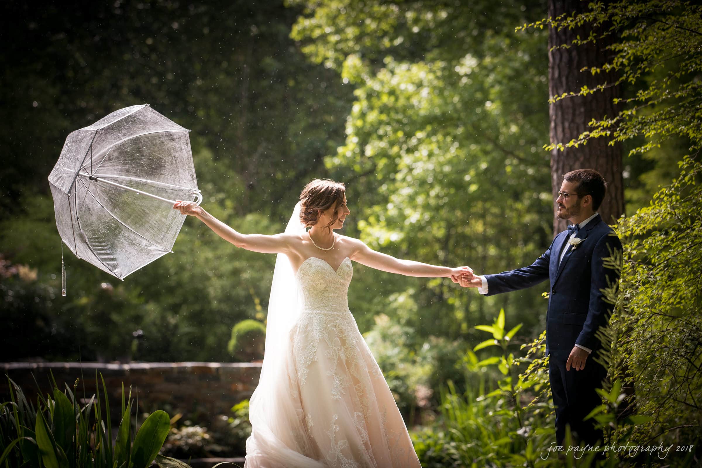 A bride in a white wedding dress holds a transparent umbrella, smiling and reaching out to a groom in a navy suit. They stand in a lush garden with sunlight filtering through the trees.