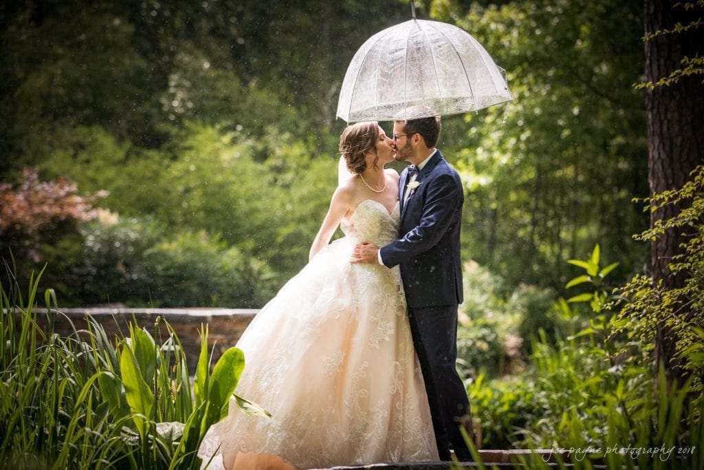 A bride and groom share a kiss under a transparent umbrella in a lush garden setting. The bride wears a flowing white gown, and the groom is in a dark suit. Sunlight filters through the trees, highlighting the raindrops around them.