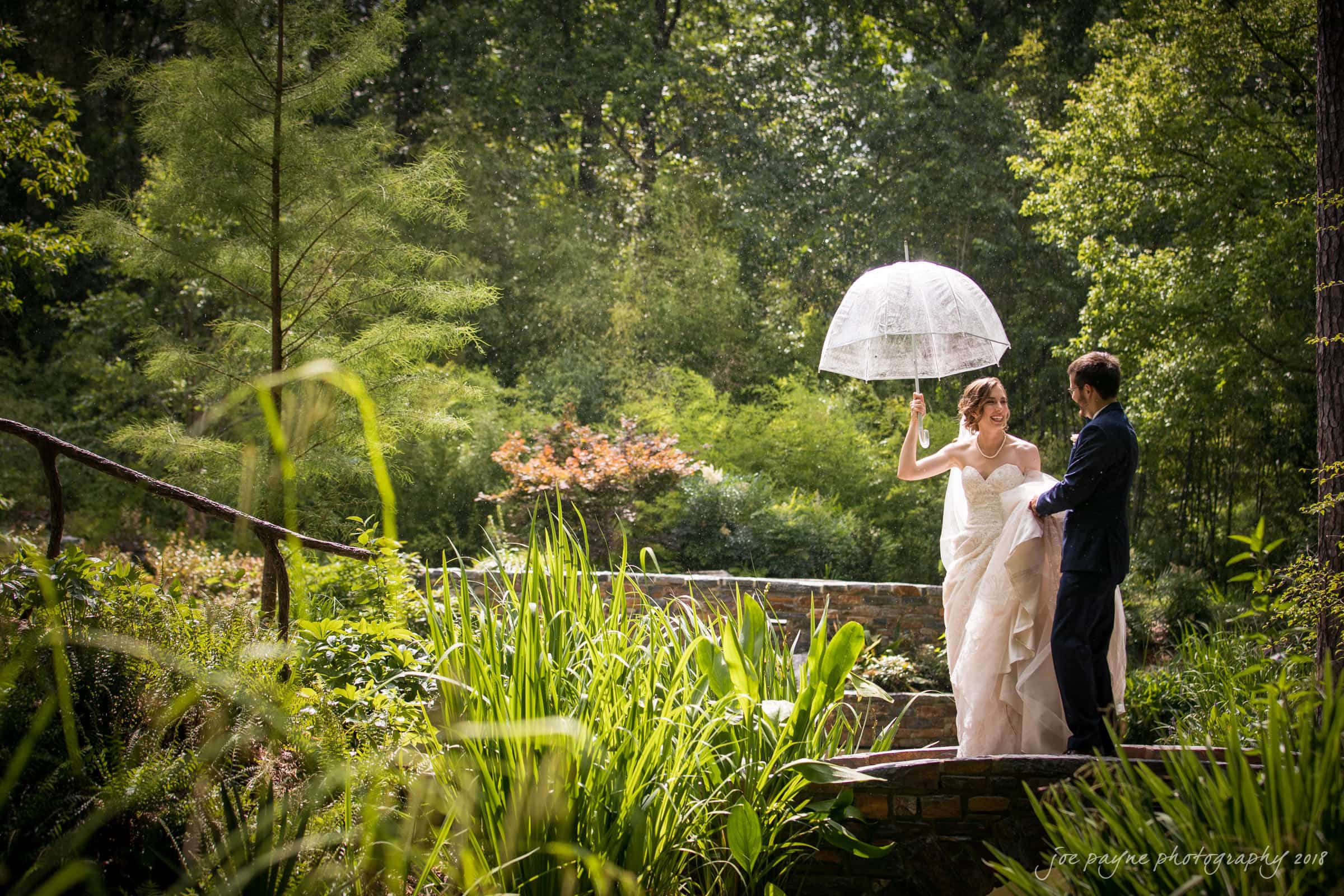 A bride and groom stand on a small bridge in a lush garden. The bride holds a lace parasol, wearing a white dress, while the groom wears a dark suit. They are surrounded by greenery and sunlight filters through the trees.