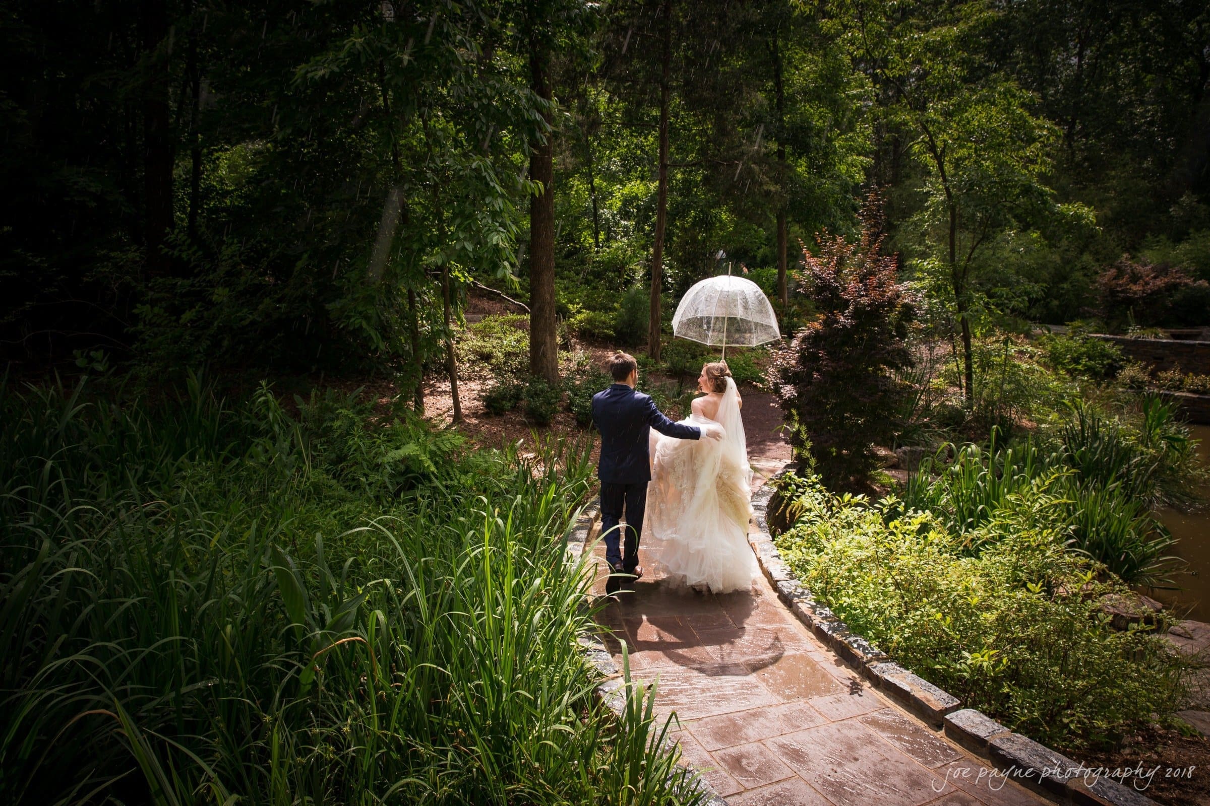 A bride and groom walk hand in hand along a stone pathway in a lush garden. The bride holds a transparent umbrella. Sunlight filters through the trees, casting dappled shadows on the path.