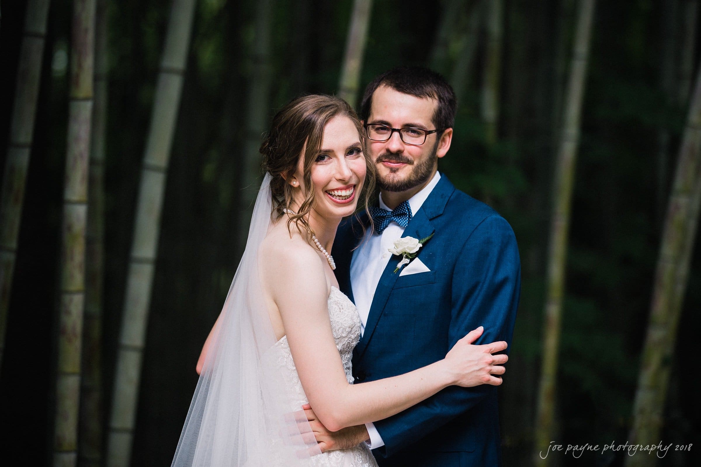 A bride in a white dress and veil embraces a groom in a blue suit and bow tie, both smiling. They are standing in front of tall bamboo, creating a serene and natural backdrop.