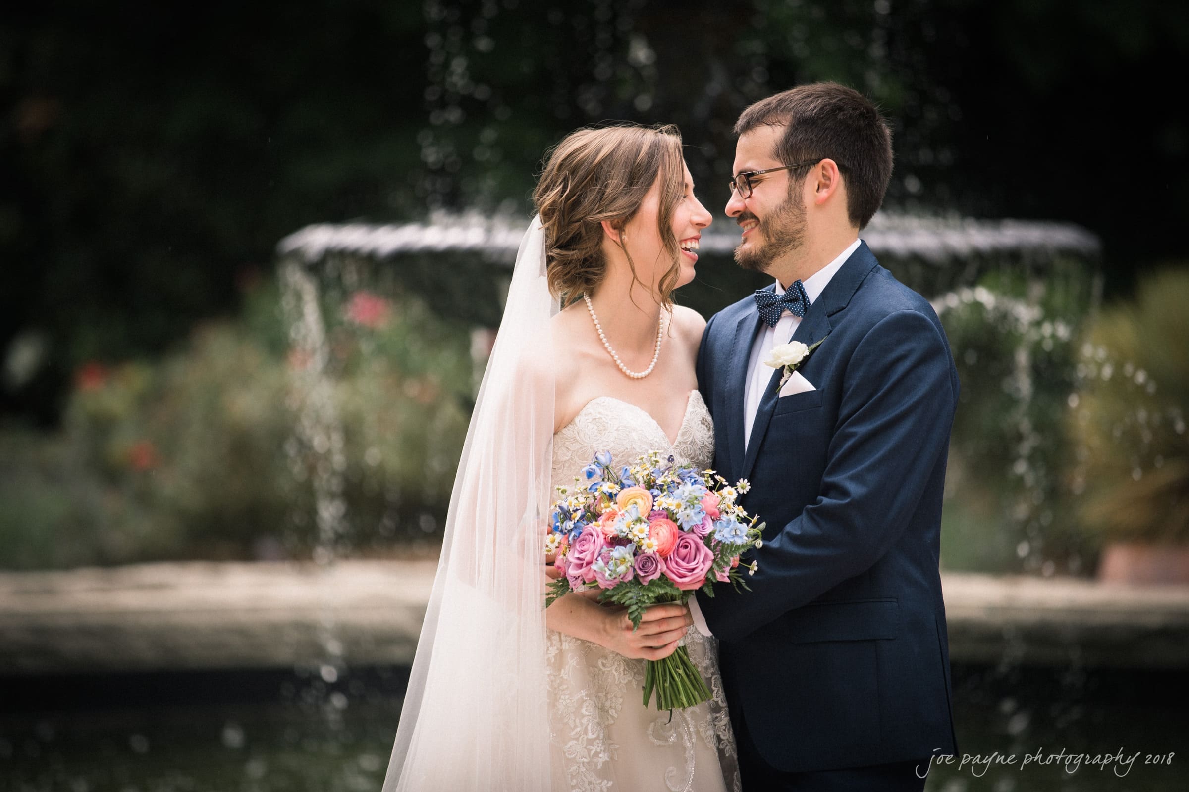 A happy couple, the bride in a white dress holding a colorful bouquet, and the groom in a blue suit, share a joyful moment in front of a fountain. Theyre looking at each other and smiling in an outdoor setting.