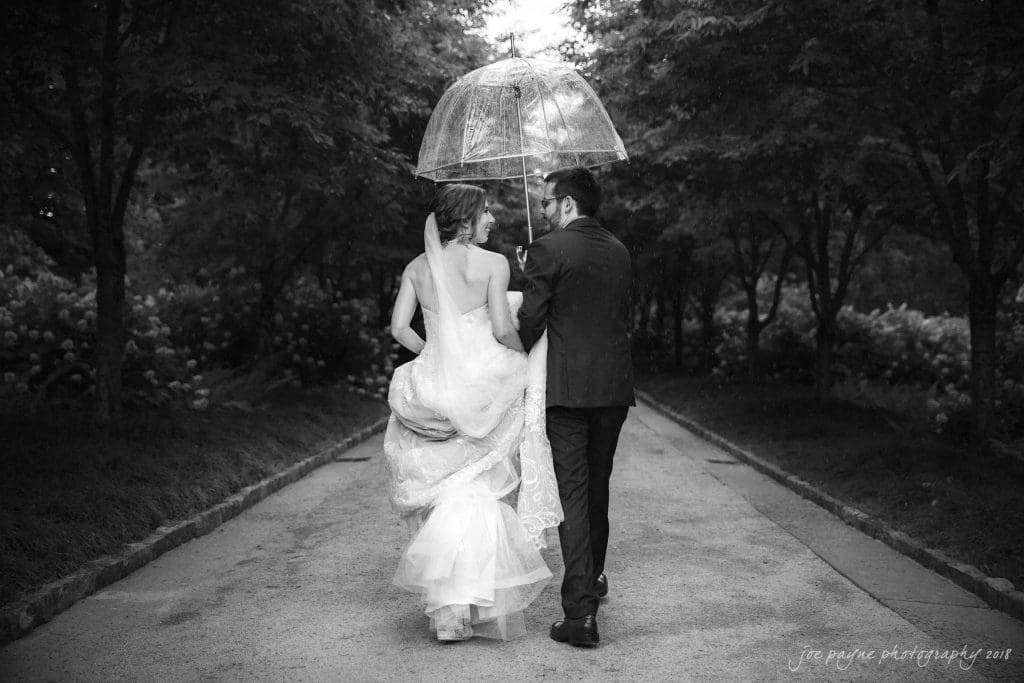 A bride and groom walk down a tree-lined path under a clear umbrella. The bride wears a long white gown, and the groom is in a dark suit. The scene is captured in black and white.