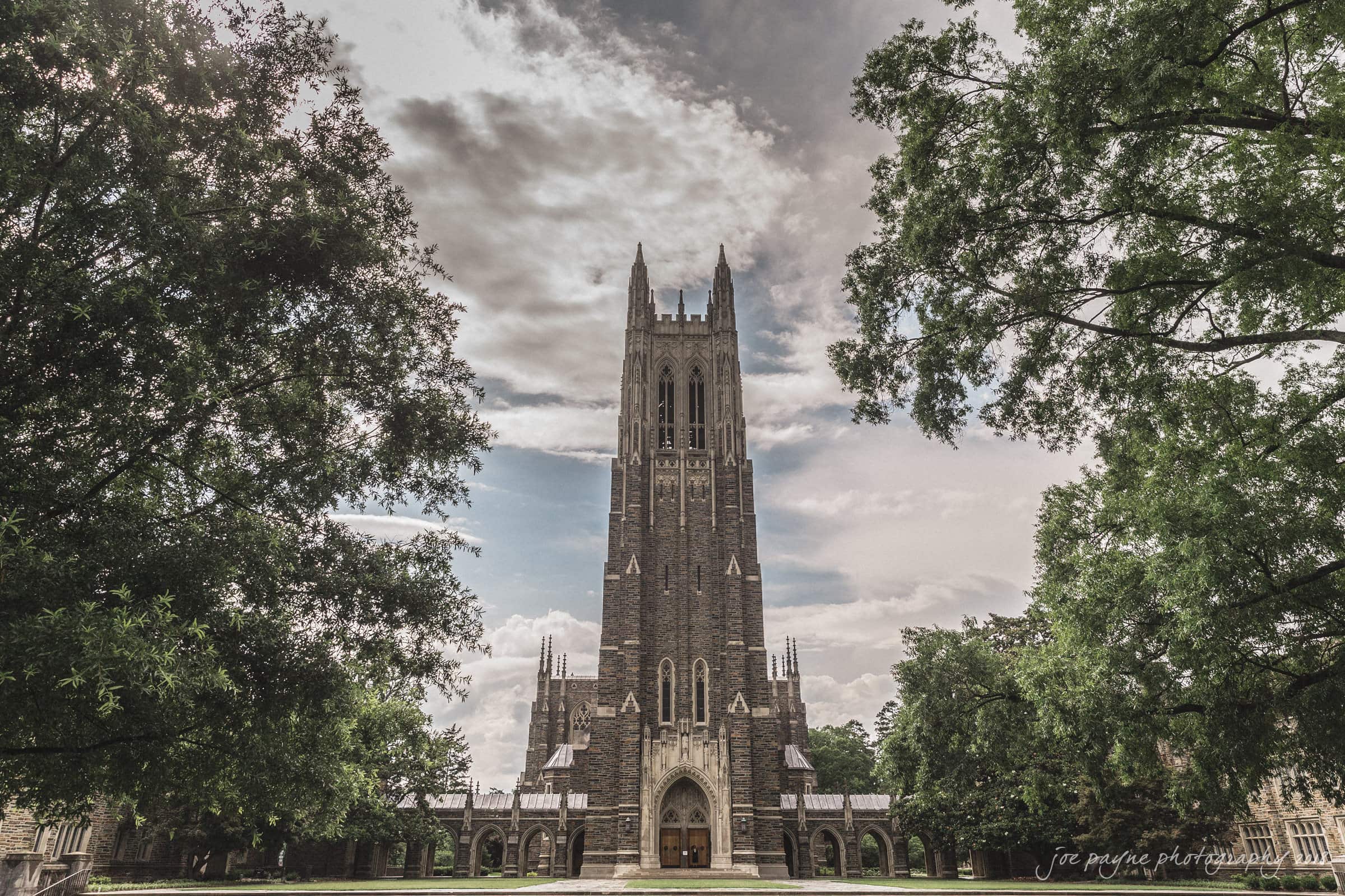 A tall, majestic Gothic-style chapel with a central spire under a cloudy sky. Surrounded by lush green trees, the structure has a dramatic and serene presence. The architecture is detailed and intricate.