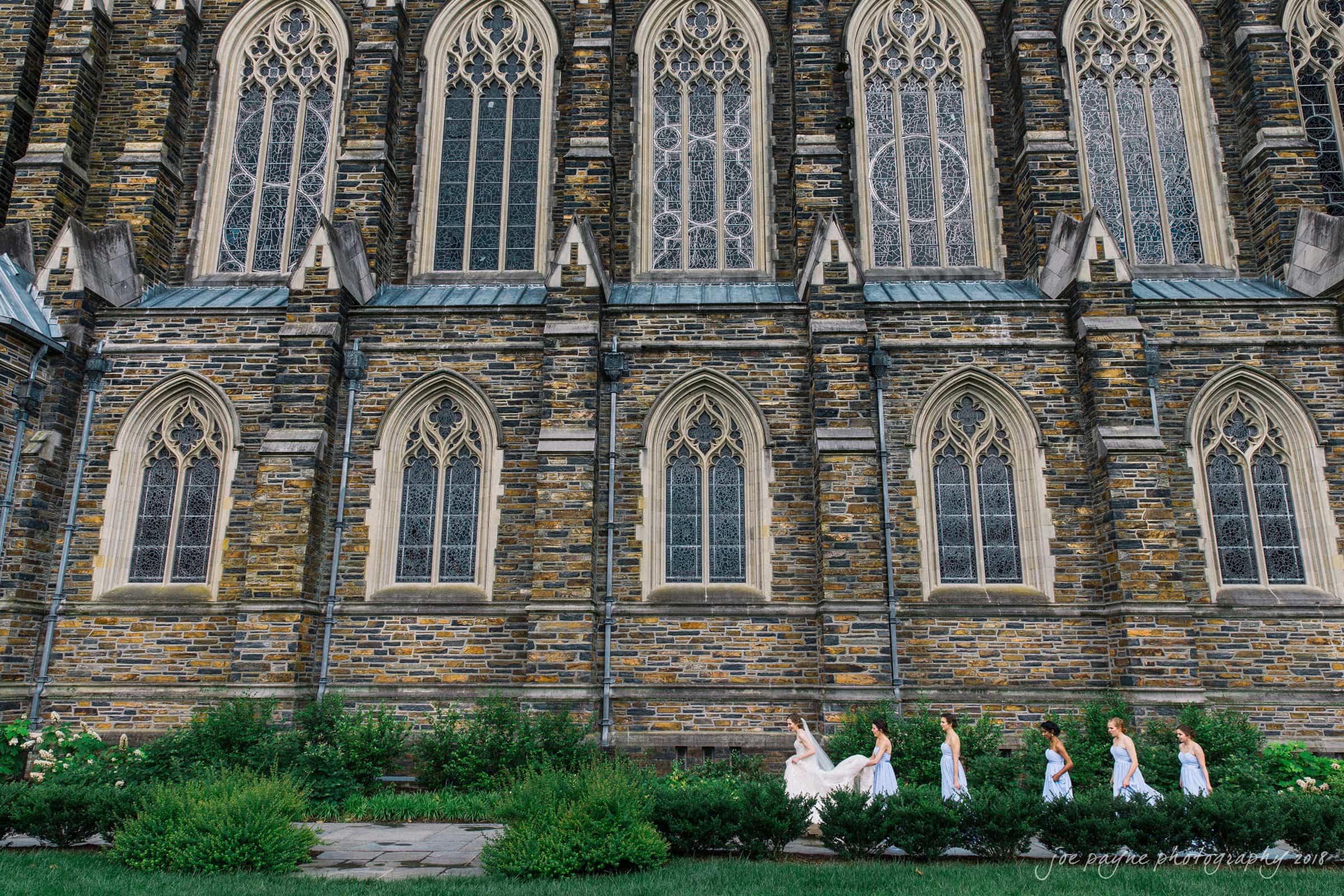 Duke Chapel Weddings A wide architectural wedding photograph at Duke Chapel, showing the bride and her bridesmaids walking in profile along the exterior stone walls beneath the towering gothic stained-glass windows.