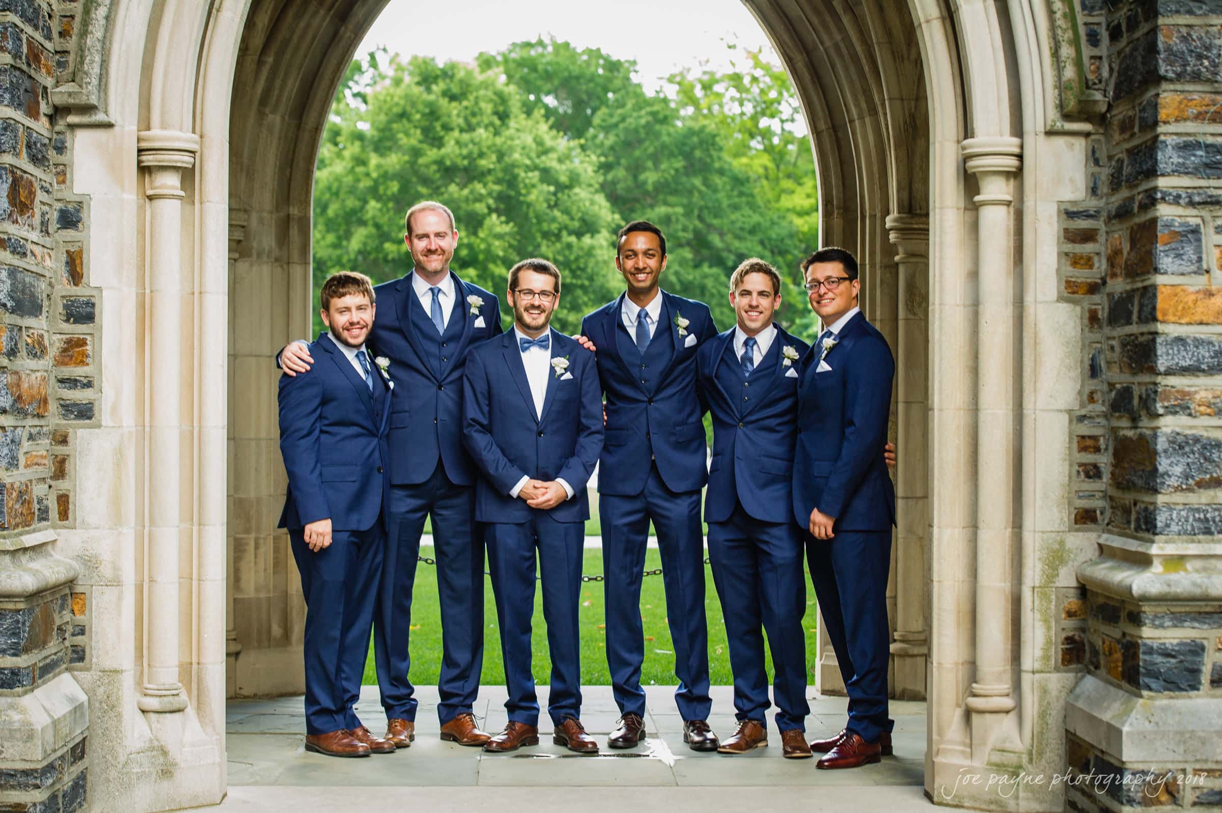 Six men in matching blue suits stand together under a stone archway. They are smiling and have their arms around each other. Green trees are visible in the background.