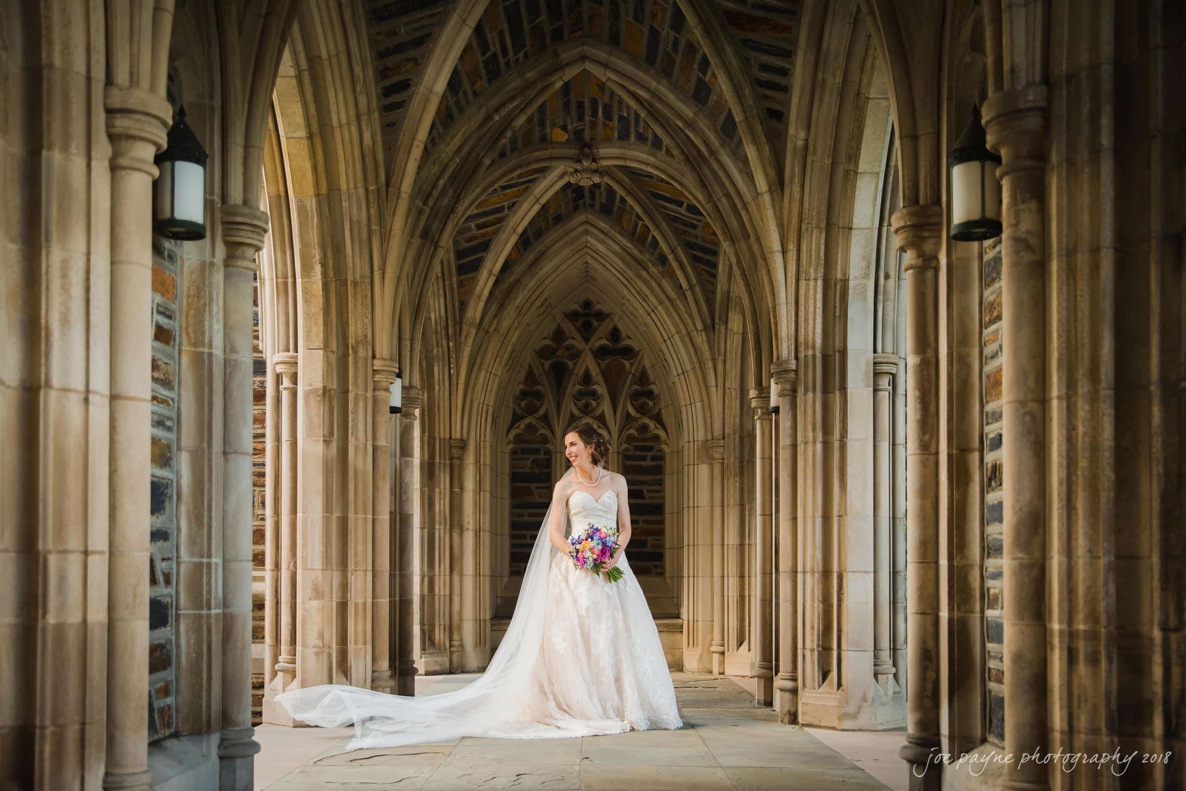 A bride in an elegant white dress stands in a gothic-style stone archway. She holds a bouquet of colorful flowers and gazes towards the side. The arches create a dramatic and romantic backdrop. Light streams in, highlighting her dress and veil.