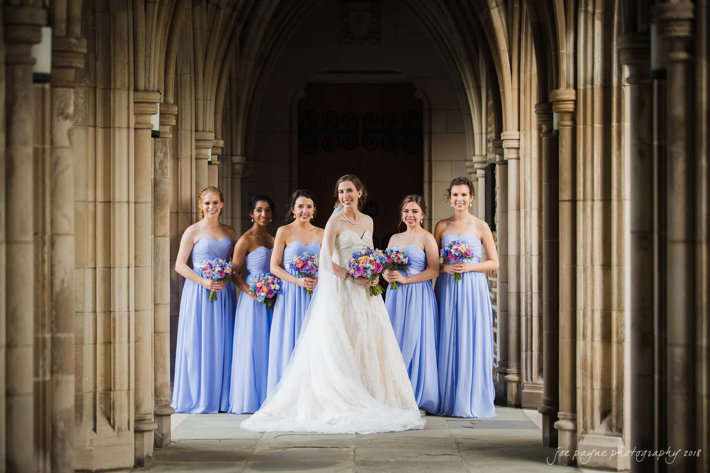 A bride in a white dress stands with five bridesmaids in light blue dresses, holding colorful bouquets. They pose under an arched stone walkway, creating a picturesque and elegant scene.