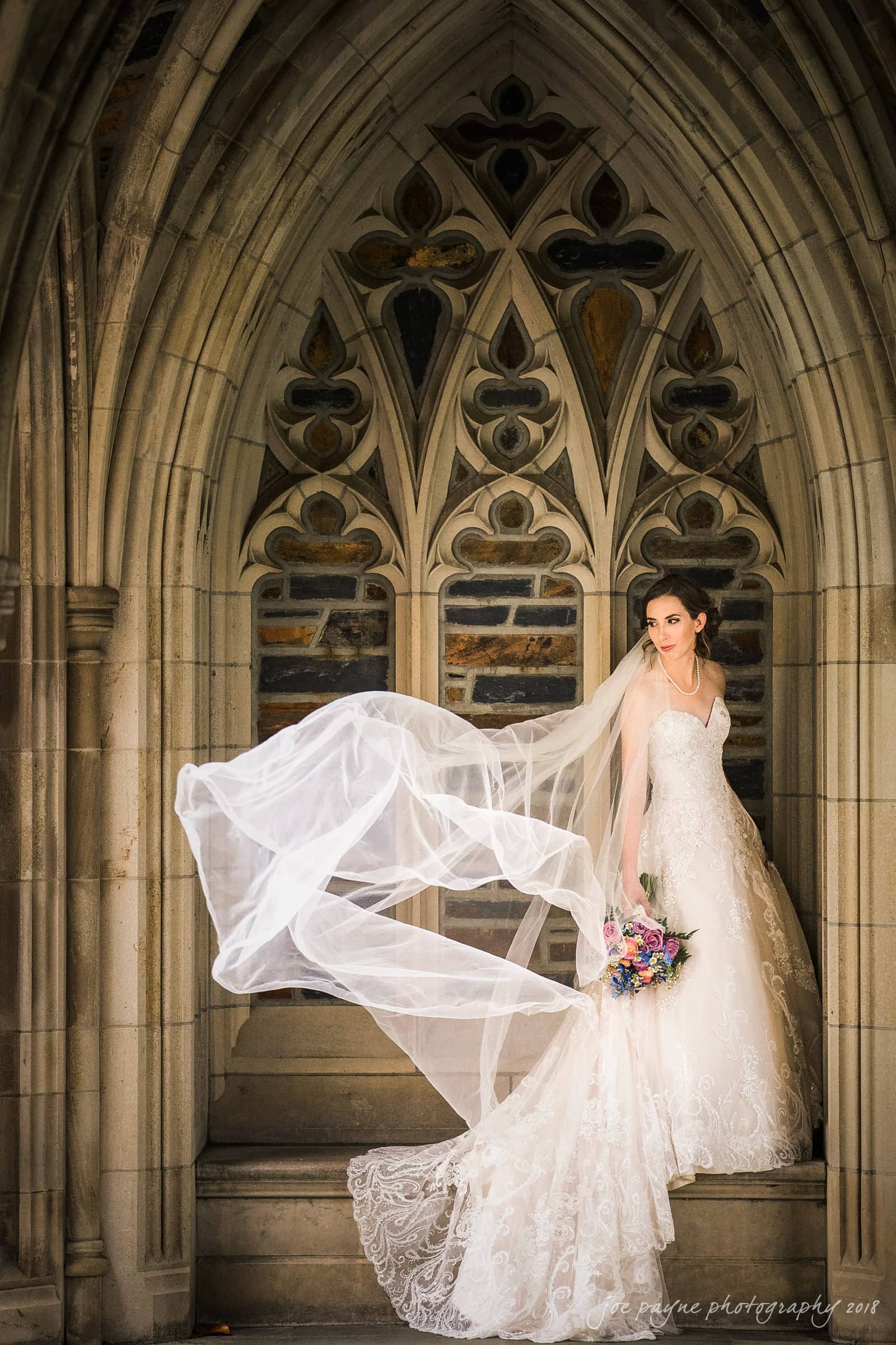 A bride stands gracefully on a stone staircase beneath a Gothic-style archway. Her dress is embellished with lace, and a long veil flows dramatically in the breeze. She holds a colorful bouquet of flowers.